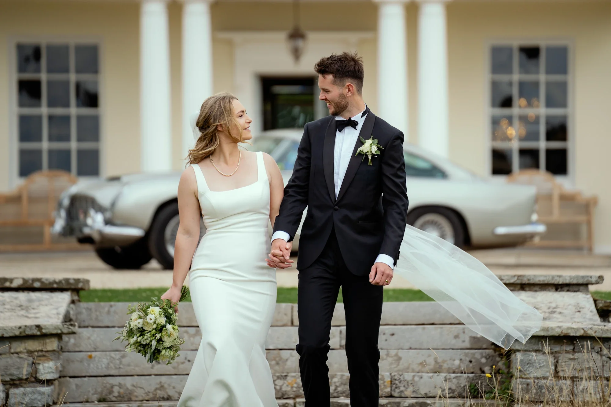 Bride and groom walking hand in hand outside a yellow house with white columns, a vintage car in the background, the bride holding a bouquet, smiling at each other.