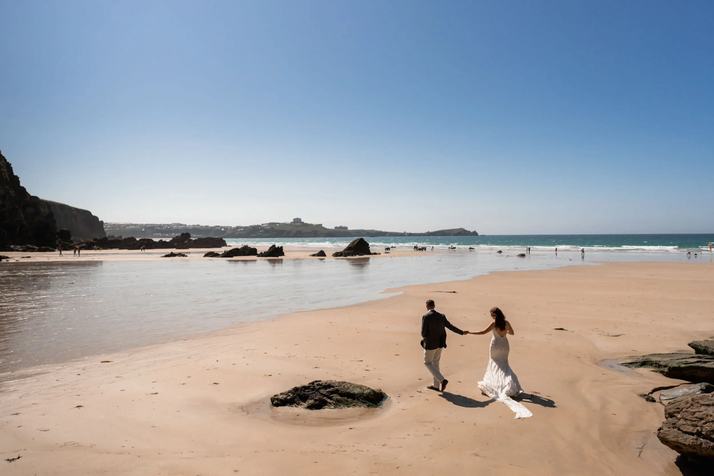 A couple holding hands walking on a sandy beach with rocks, near the shoreline under a clear blue sky.