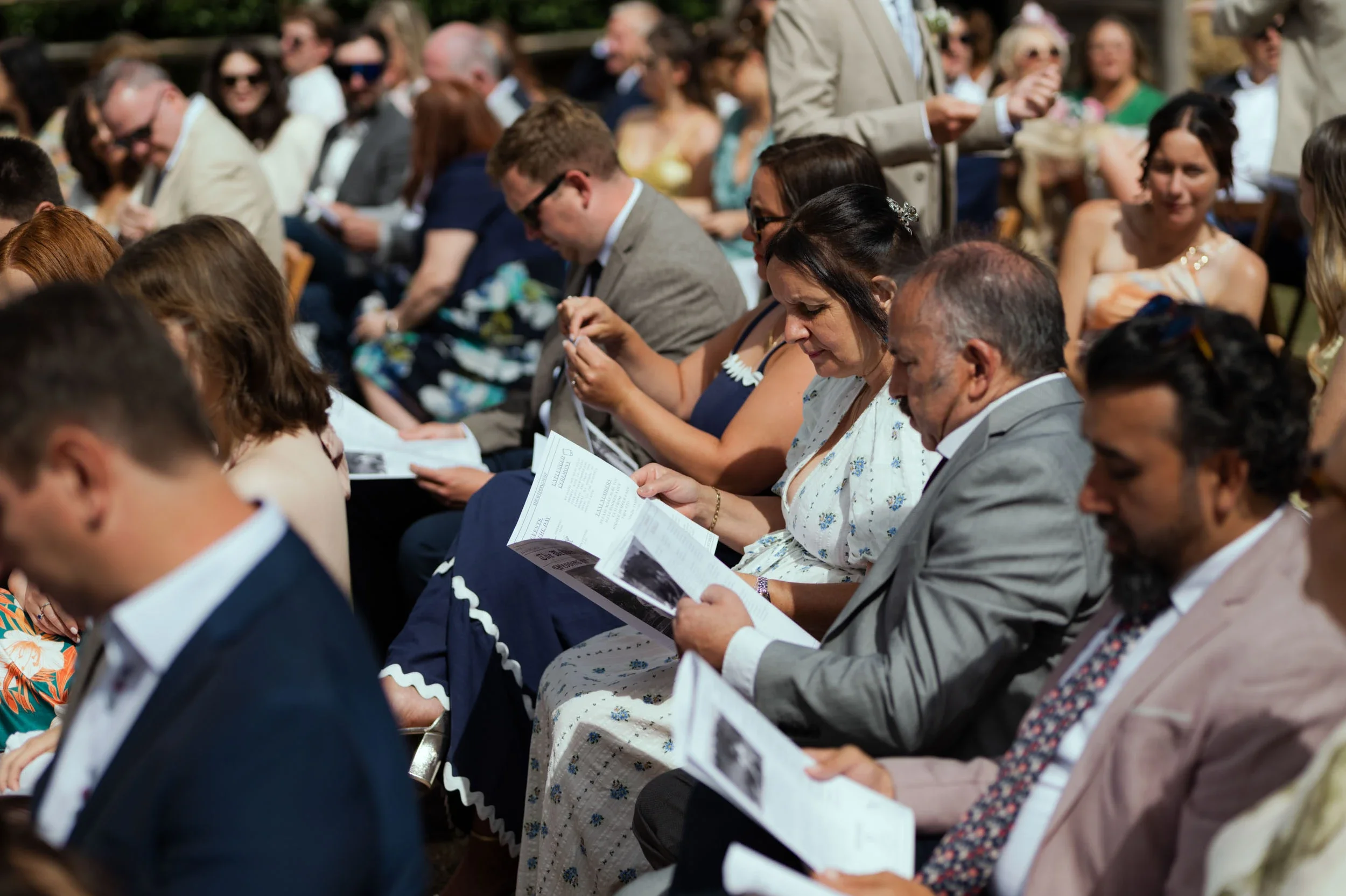 People dressed in formal attire sitting outdoors at a wedding ceremony, reading programs.