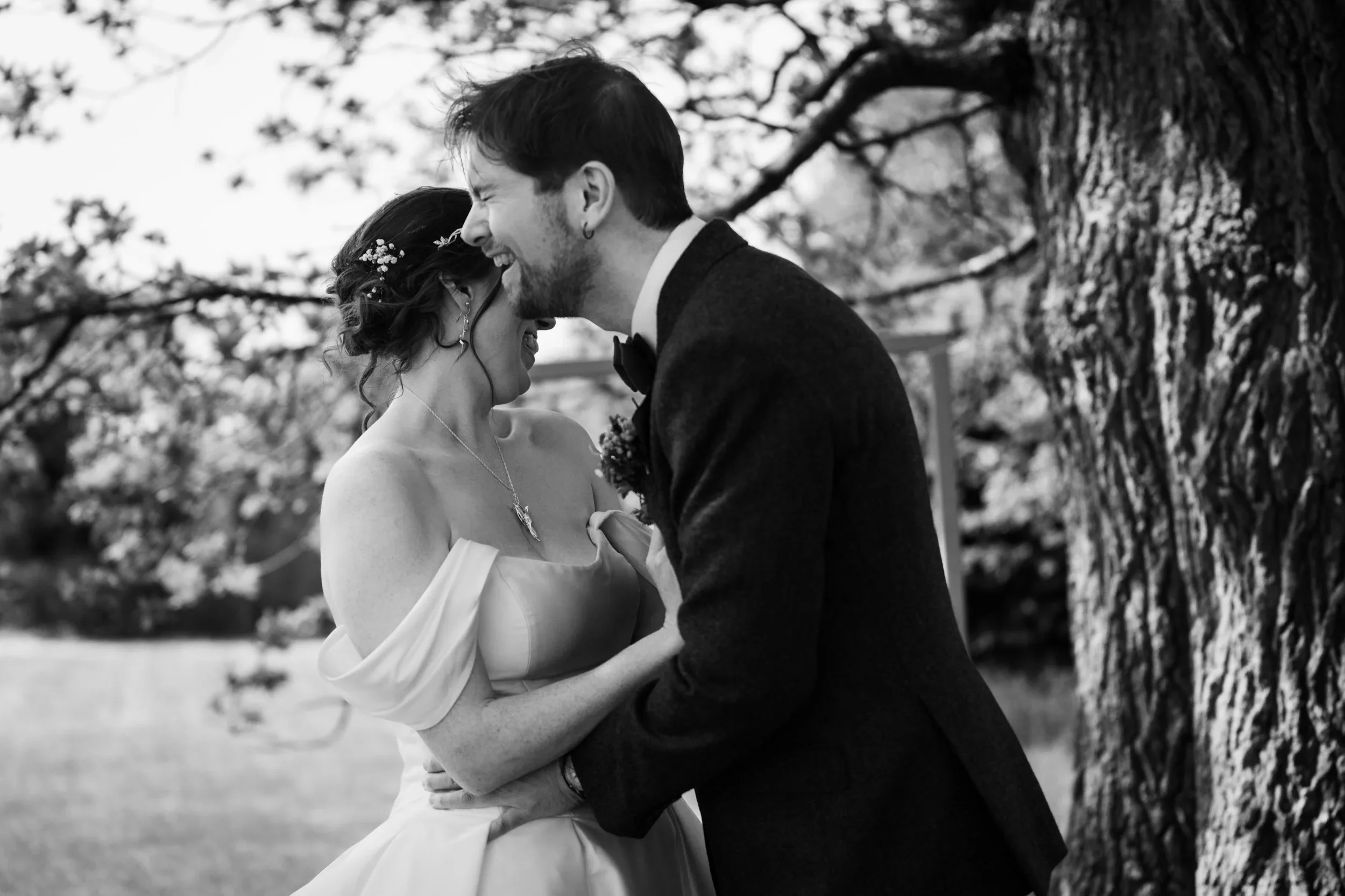 A joyful couple sharing a kiss outdoors, with a large tree in the background, during their wedding celebration.