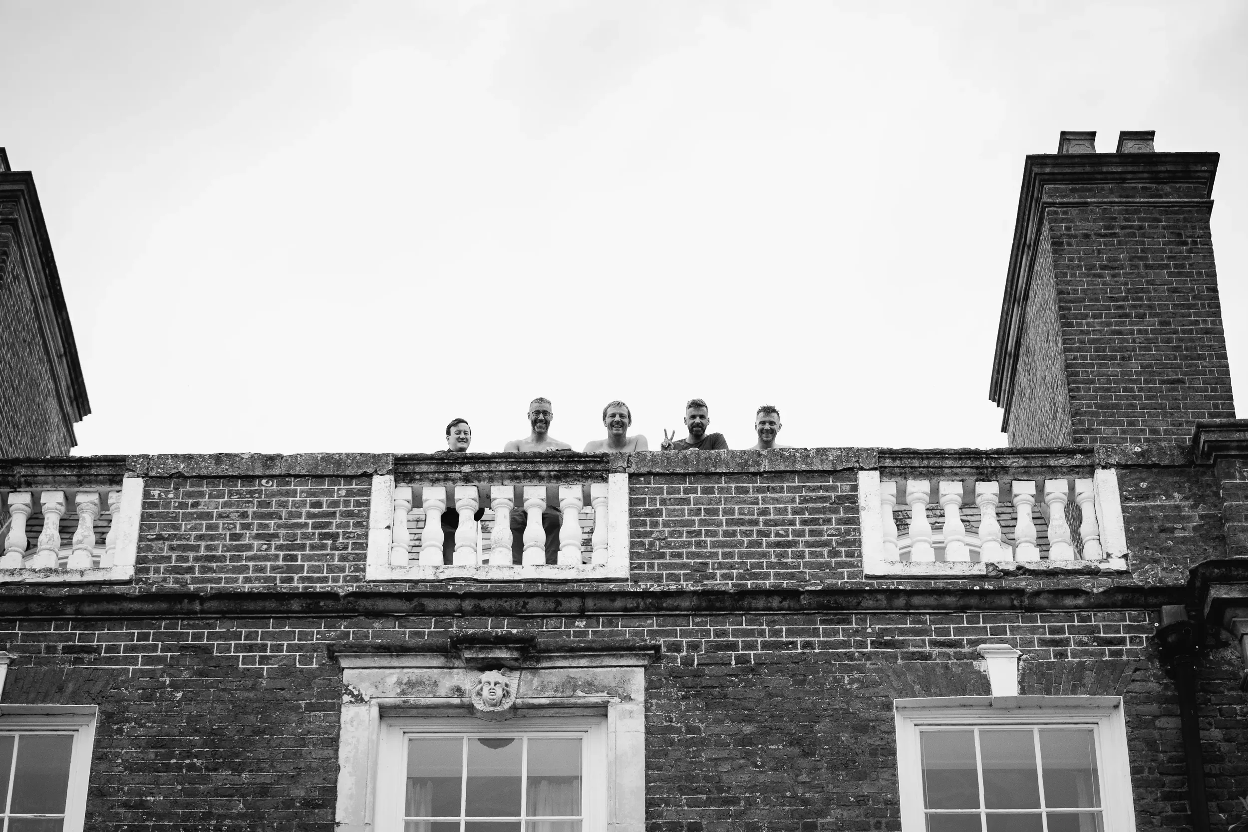 Five people are leaning over a balcony railing on the top floor of a brick building, smiling and one making a peace sign, with an overcast sky in the background.