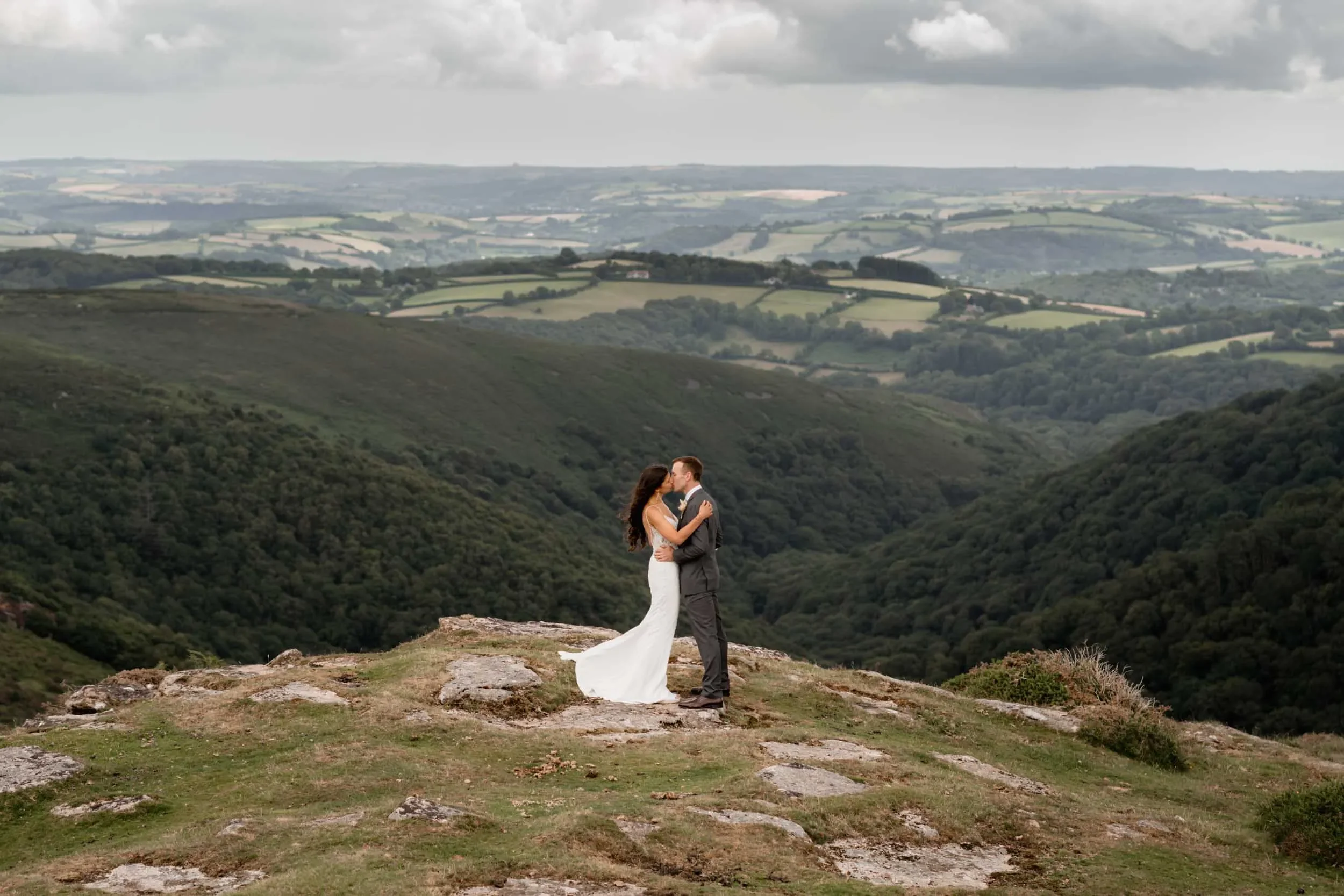 A bride and groom kissing on a rocky hilltop with a lush green valley and rolling hills in the background.