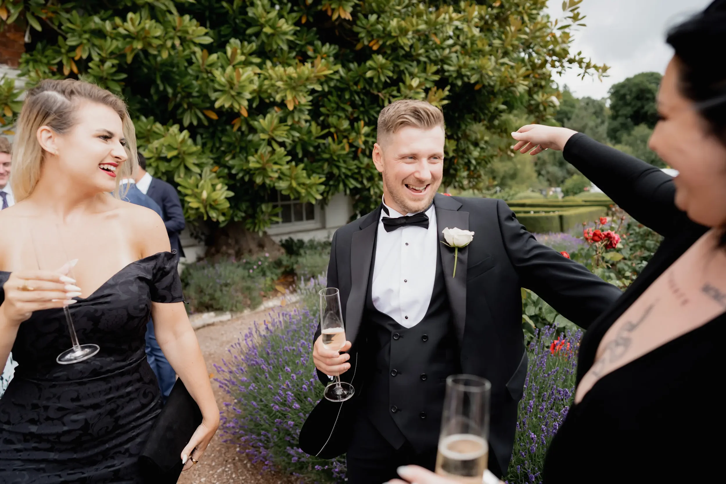 People at a wedding reception outdoors, including a man in a tuxedo and a woman with blonde hair in a black dress, smiling and holding champagne glasses, with others in the background.