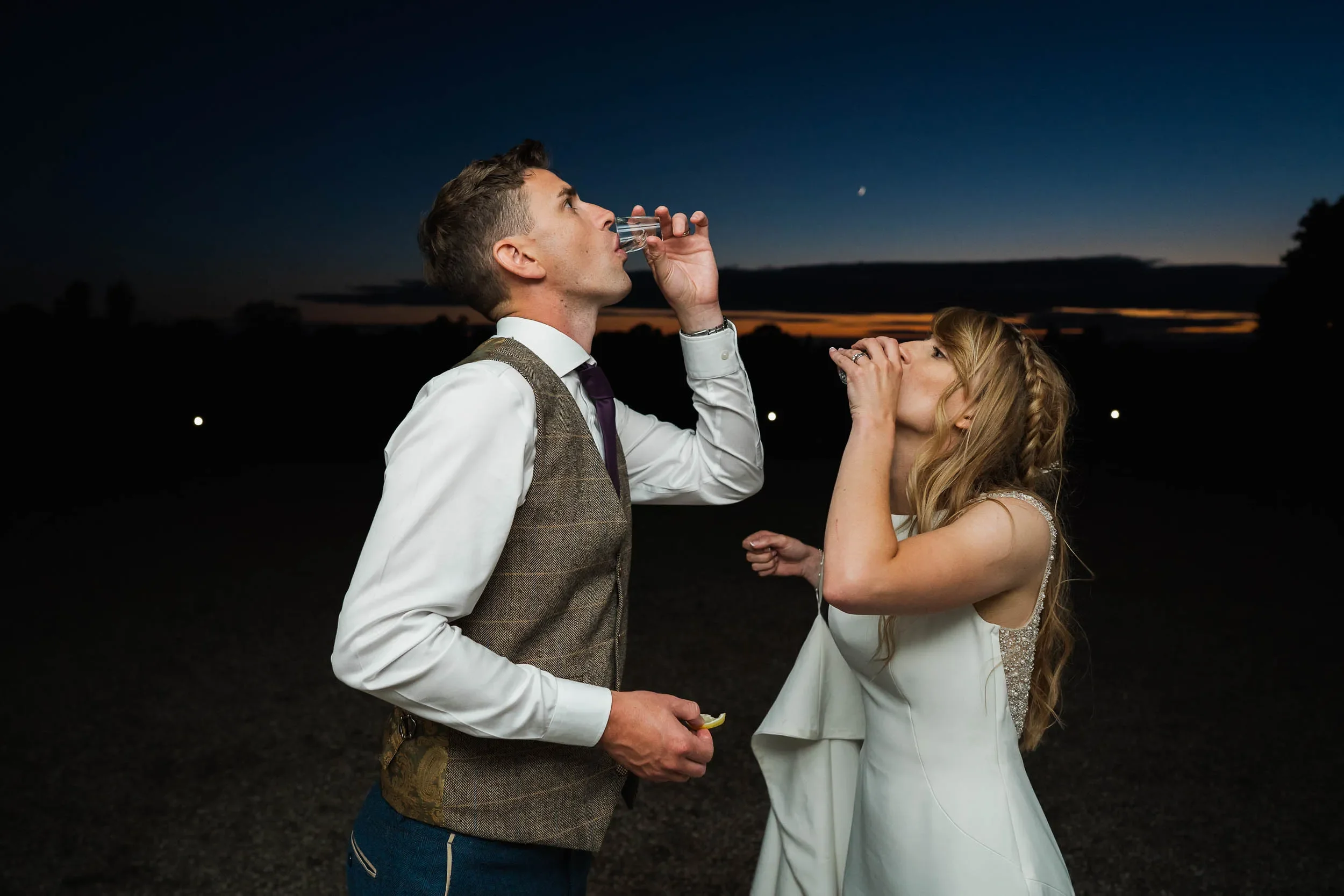 A bride and groom in wedding attire blowing shot glasses into each other's mouths outdoors at dusk.