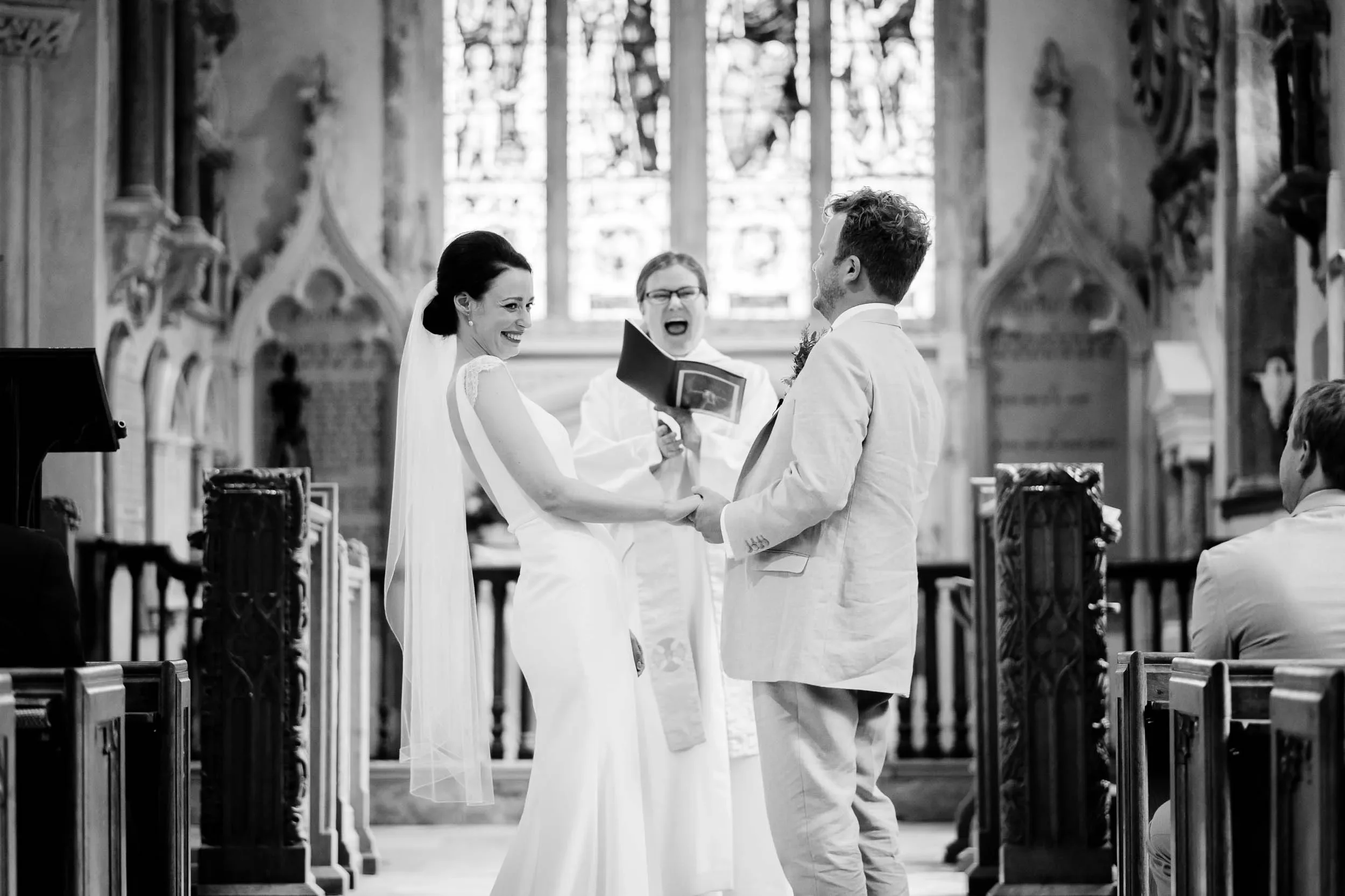A black and white photo of a wedding ceremony inside a church with a bride and groom holding hands, smiling, facing each other, with an officiant standing behind them. The bride is wearing a white dress with a veil, and the groom is in a light-colore