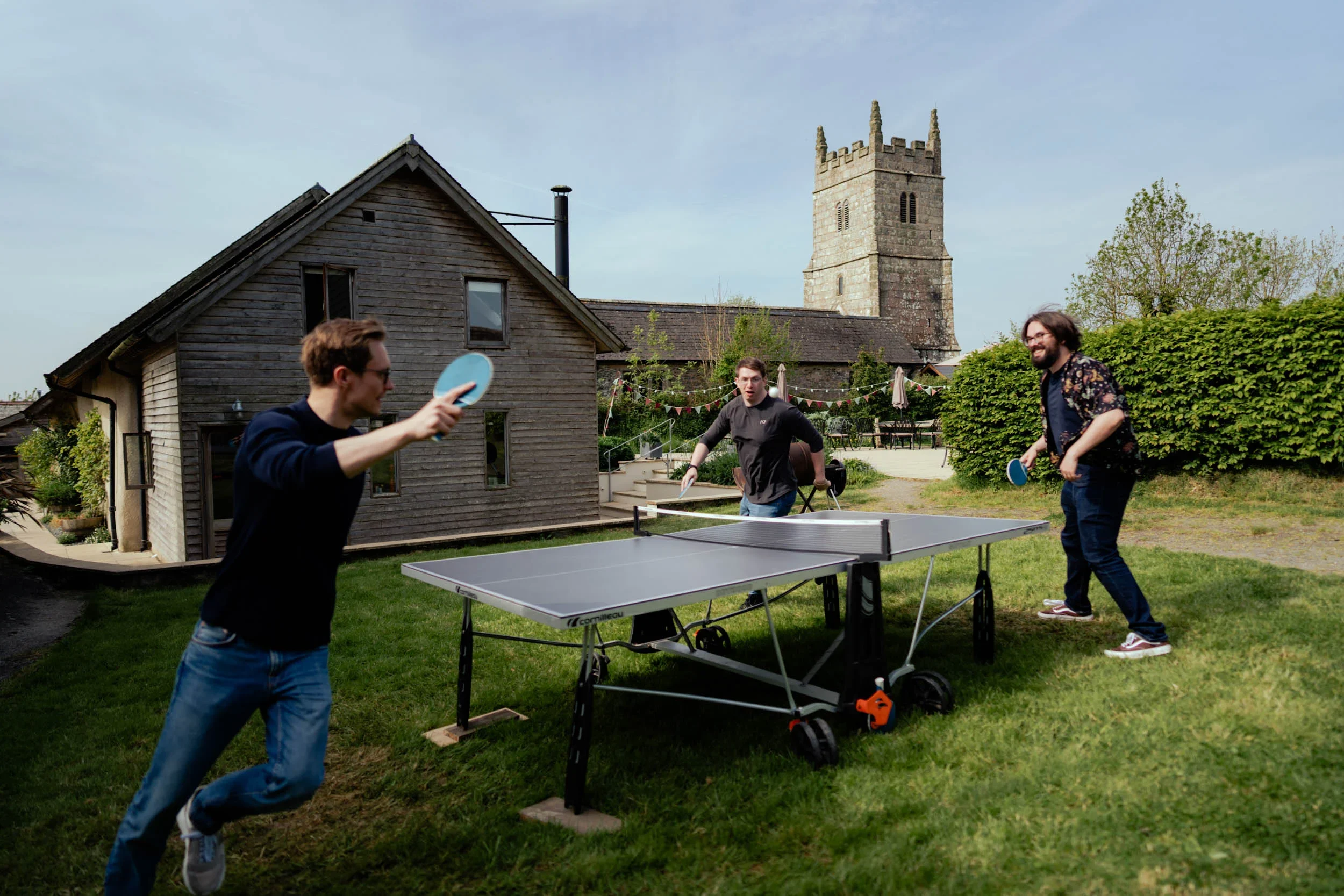 Three men playing table tennis outdoors on a sunny day near a rustic house with a church tower in the background.