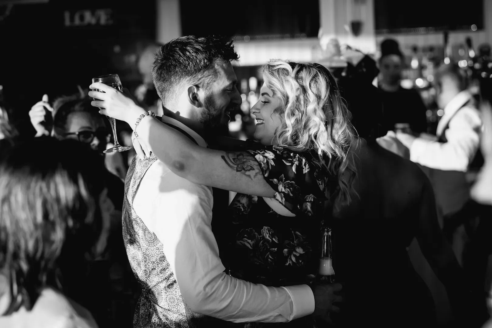 A couple embracing and smiling at each other during a celebration, with one holding a wine glass, at a lively event or party at night.