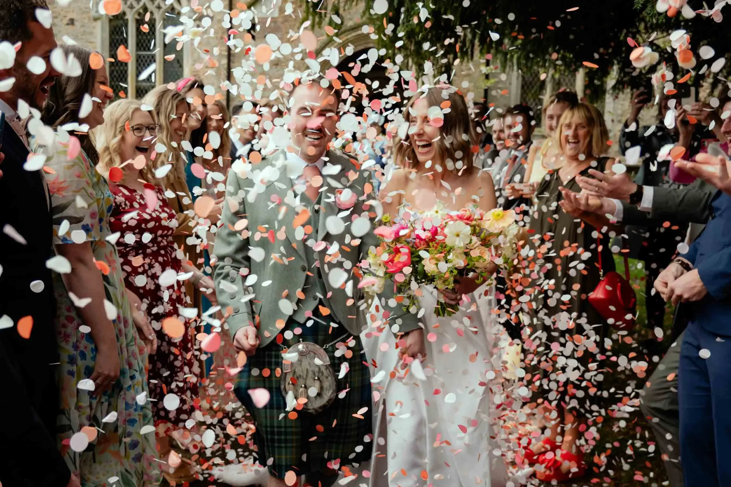 A newly married couple surrounded by friends and family, celebrating with confetti and flowers during their wedding.