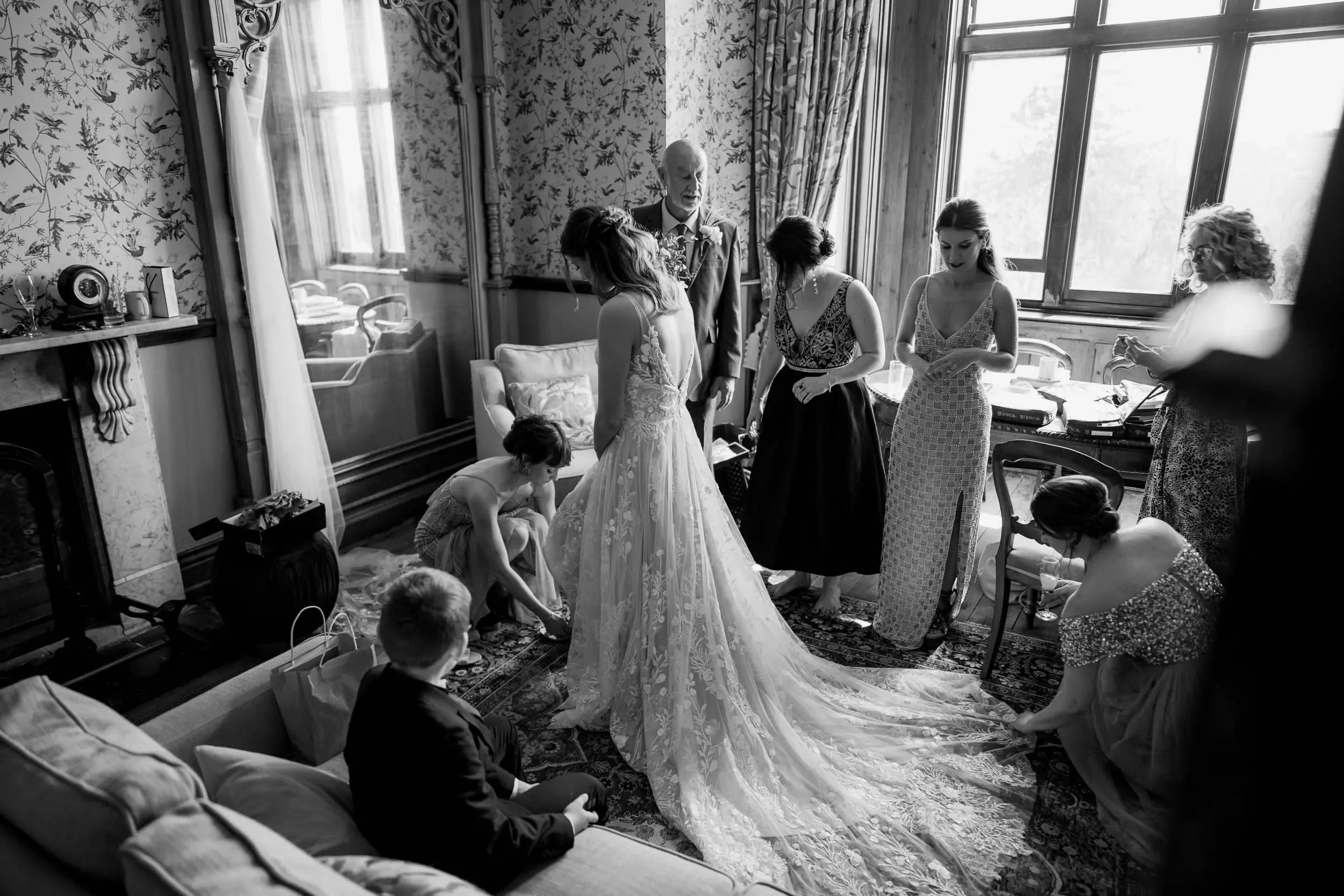 Bride in a wedding dress surrounded by bridesmaids and children, getting ready in a vintage-style room with floral wallpaper and large windows.