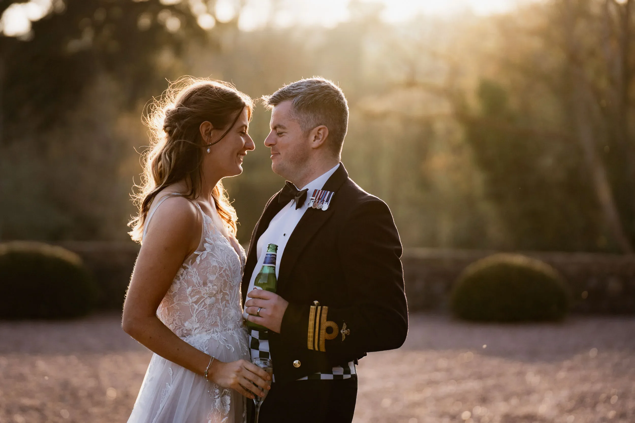 A bride and groom stand closely together outdoors at sunset, smiling and holding drinks.