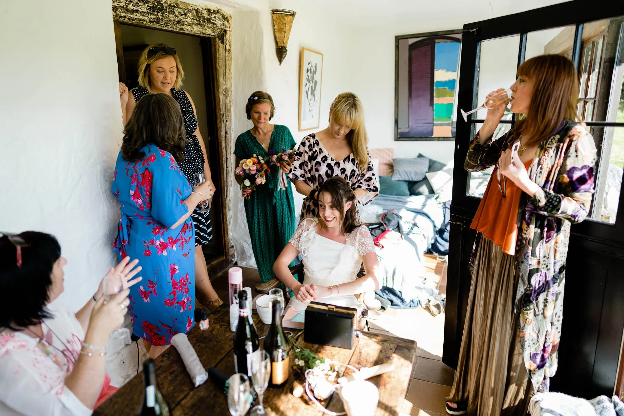 A group of women, including a bride in a white dress, gather in a cozy room with sunlight, holding drinks, flowers, and celebrating.
