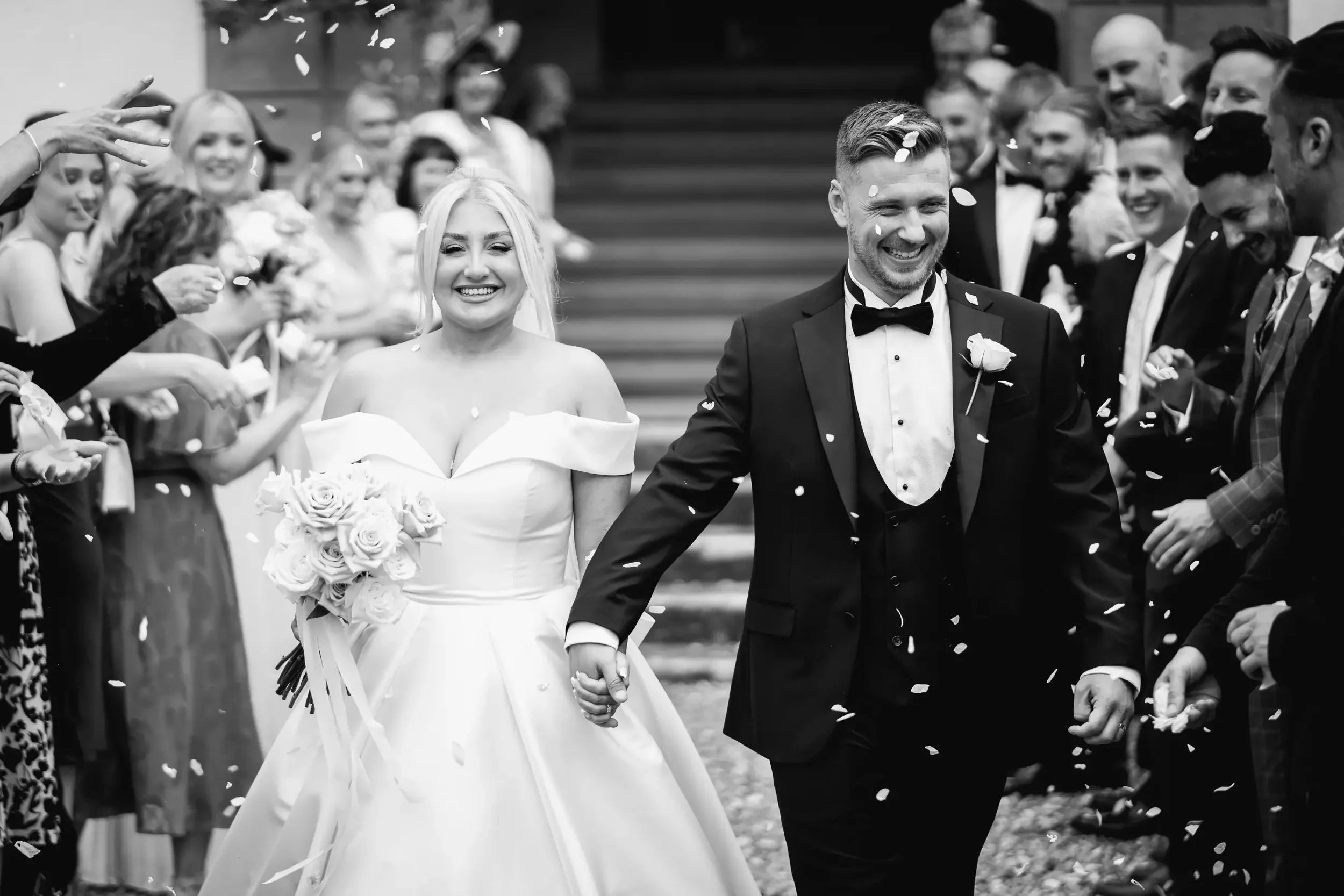 A bride and groom holding hands and smiling as they walk through a crowd of guests, throwing confetti in celebration at their wedding.