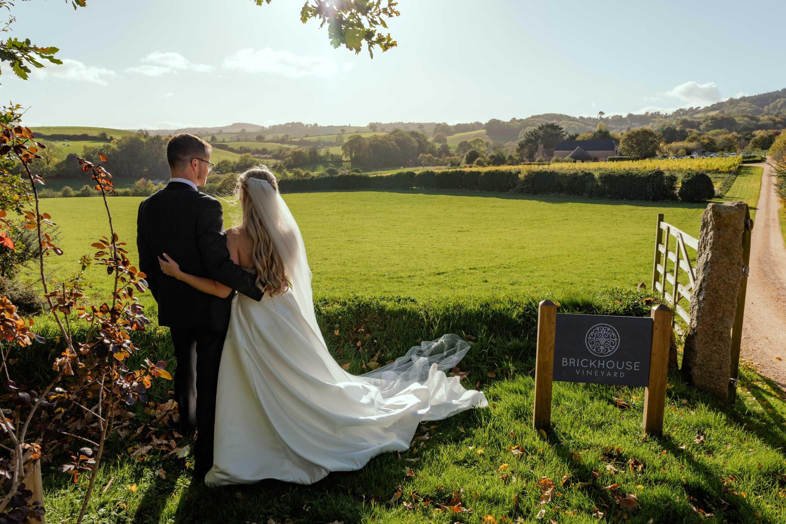 A couple in wedding attire stands together outdoors in a lush green field, with the bride wearing a white gown and veil, and the groom wearing a black suit. They are facing away from the camera, embracing in front of a sign that reads 'Brickhouse Vin