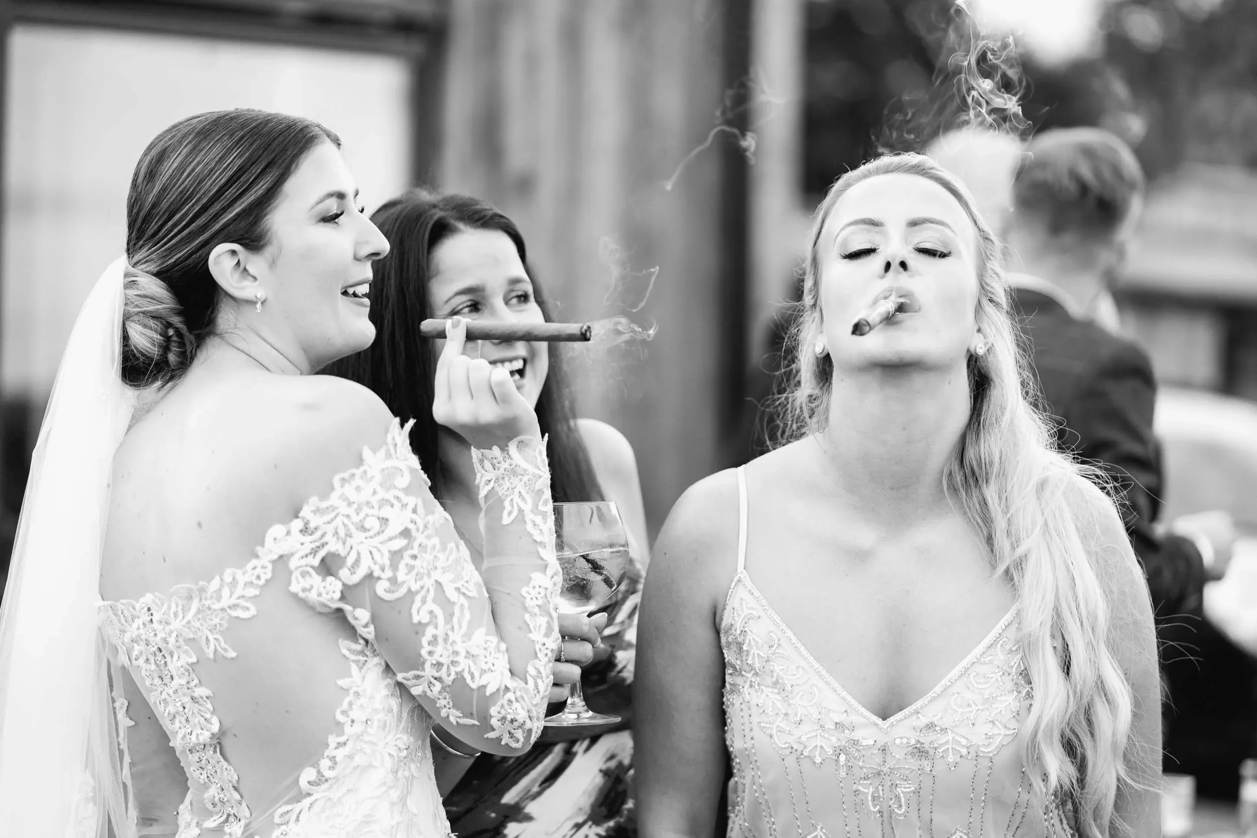 A wedding celebration with three women, one in a wedding dress, holding a cigar and a glass of wine, with a fourth woman in the background, all outdoors.