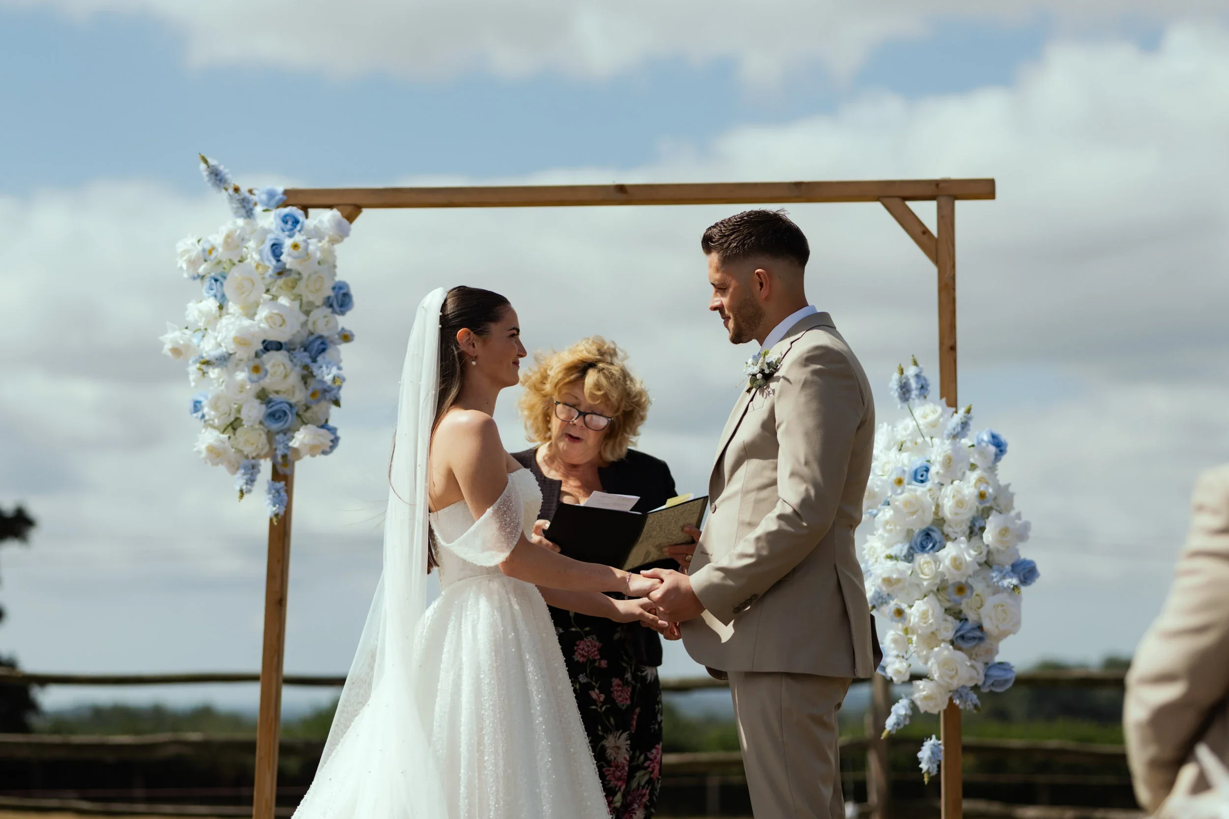 A bride and groom holding hands during their outdoor wedding ceremony with an officiant, under a wooden arch decorated with white and blue flowers, with a cloudy sky in the background.