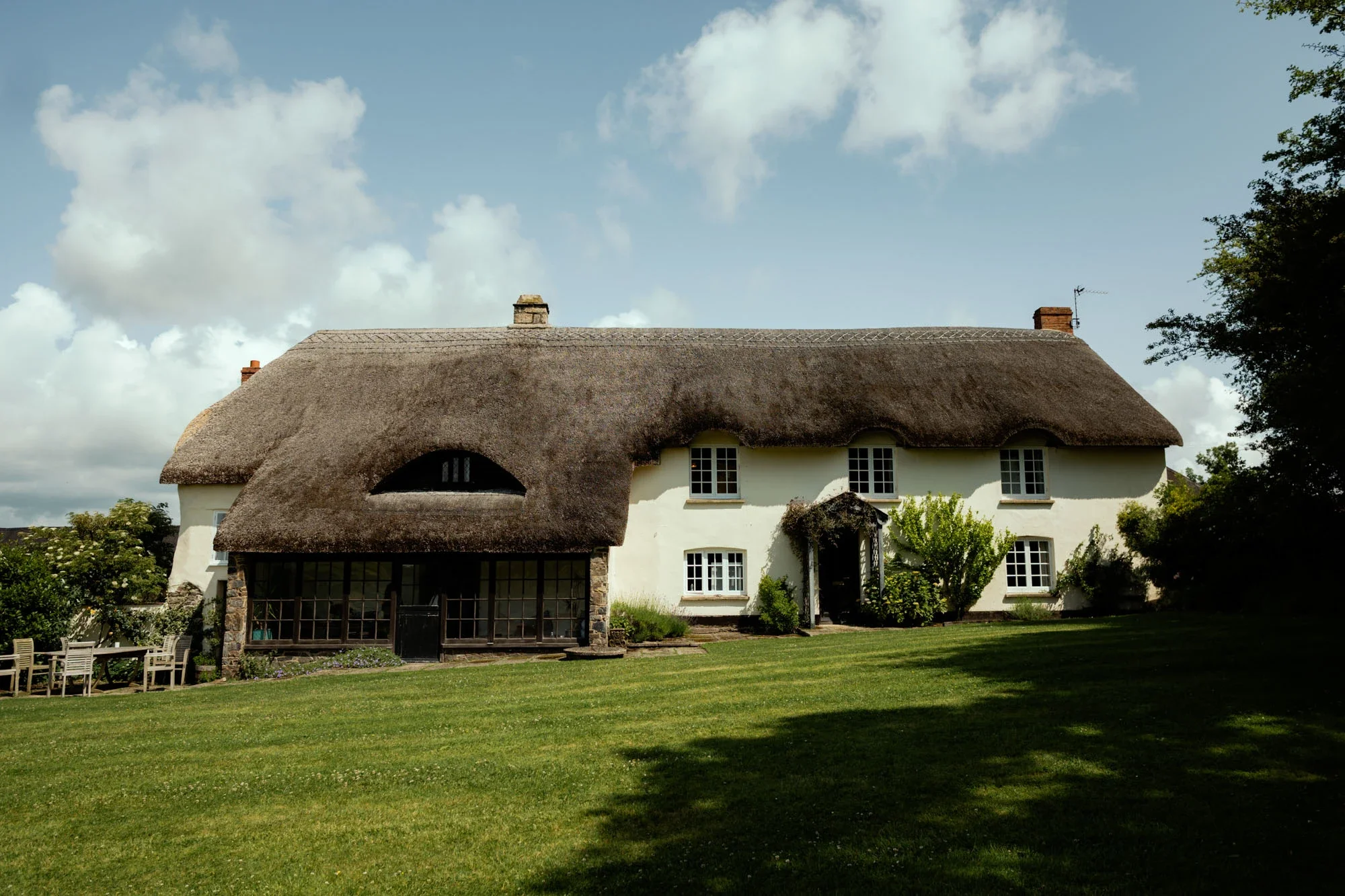 A large cottage with white walls and a thatched roof surrounded by a green lawn and trees, under a partly cloudy sky.