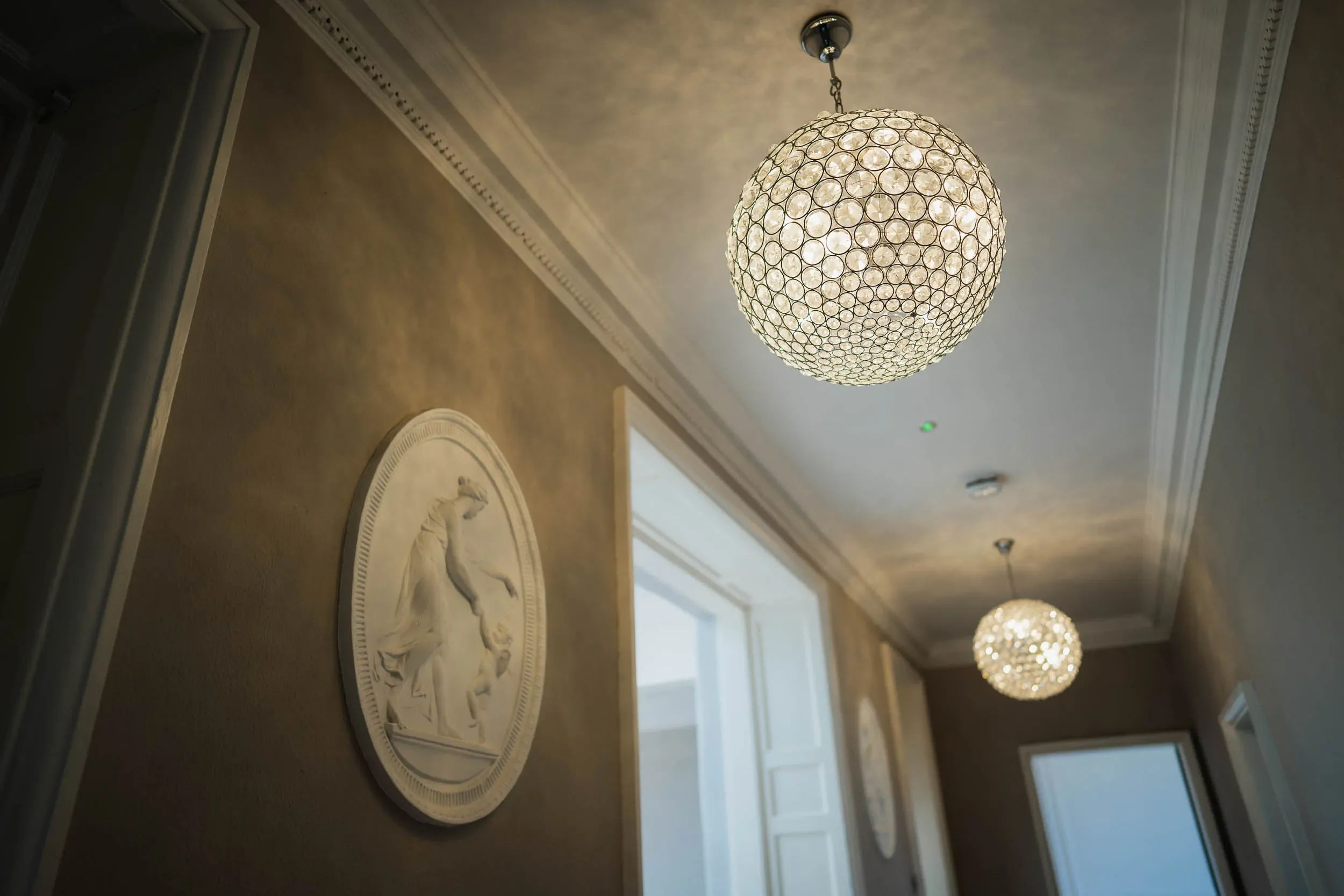 Interior view of a hallway with beige walls, ornate crown molding, two spherical chandeliers hanging from the ceiling, and framed art pieces on the wall. Natural light streams in through a window.