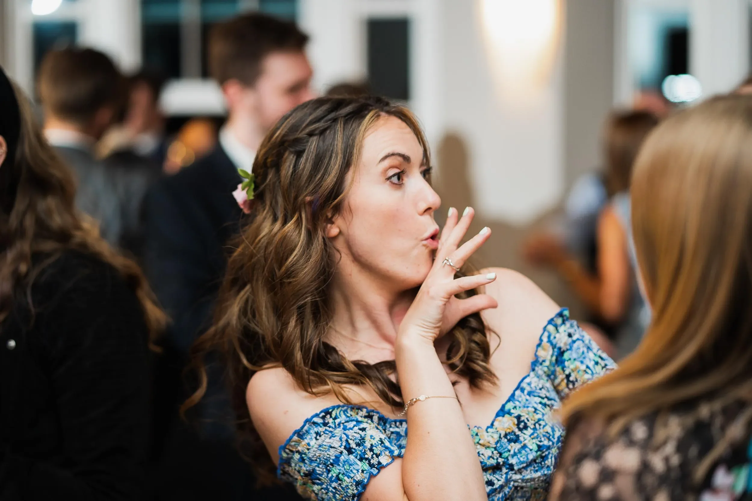 A woman with long brown hair, wearing a blue off-shoulder dress with floral patterns, is making a shushing gesture with her index finger over her lips at an indoor event. She has a flower in her hair and is surrounded by other people.