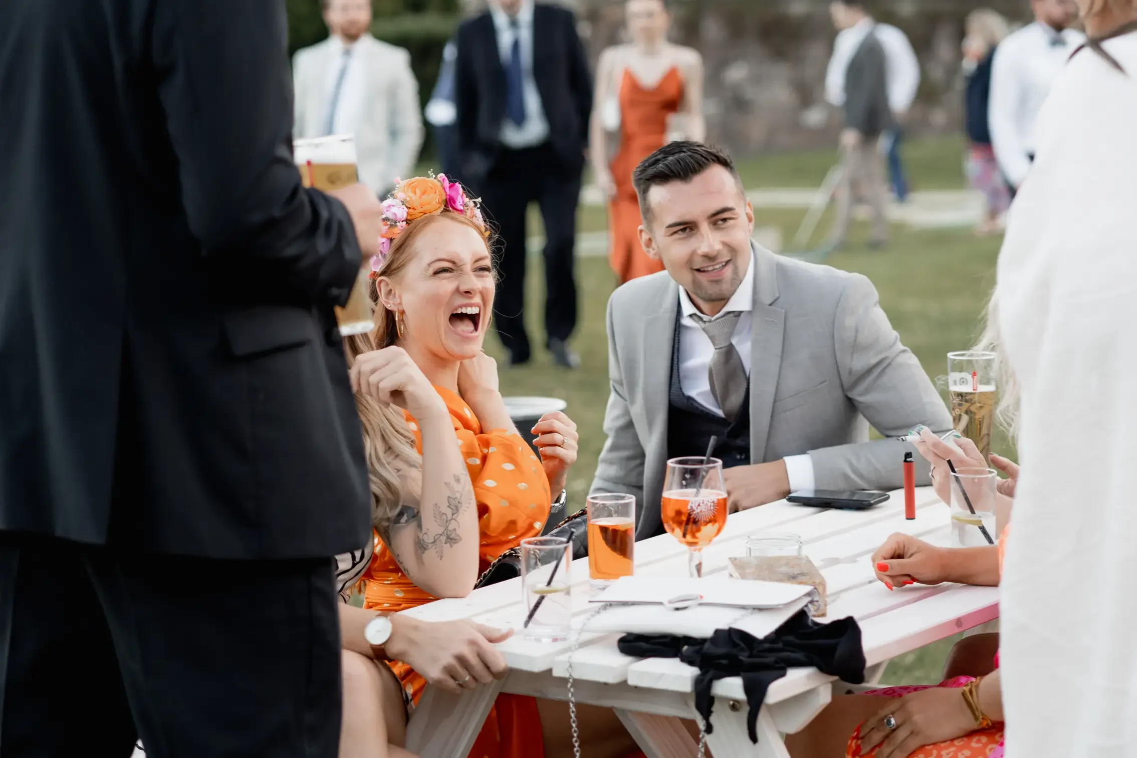 People socializing outdoors at a party, with a woman laughing and wearing a floral headband and orange dress, and a man in a grey suit smiling, seated at a white table with drinks and makeup products.