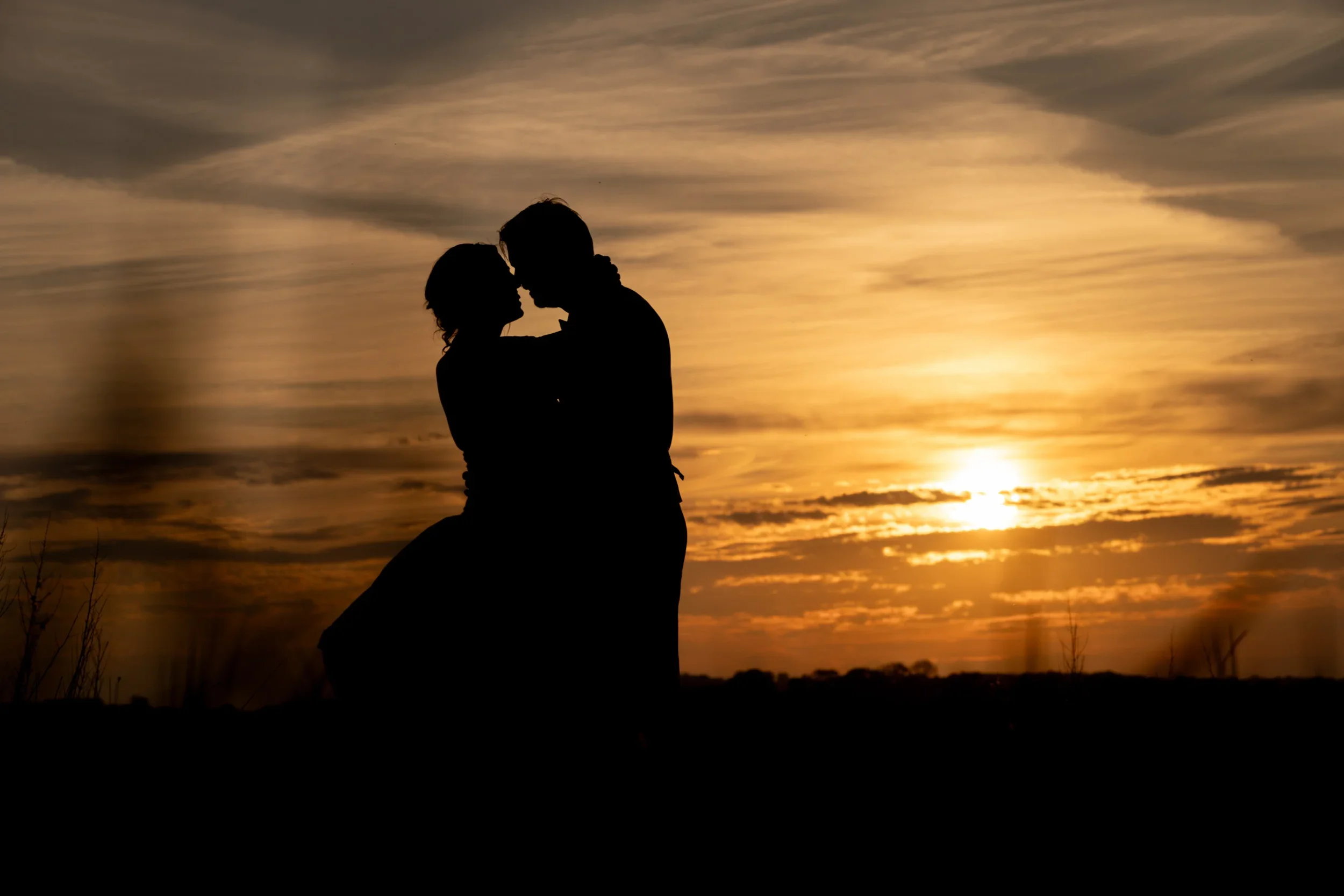 Silhouette of a couple embracing during sunset with a colorful sky and clouds in the background.