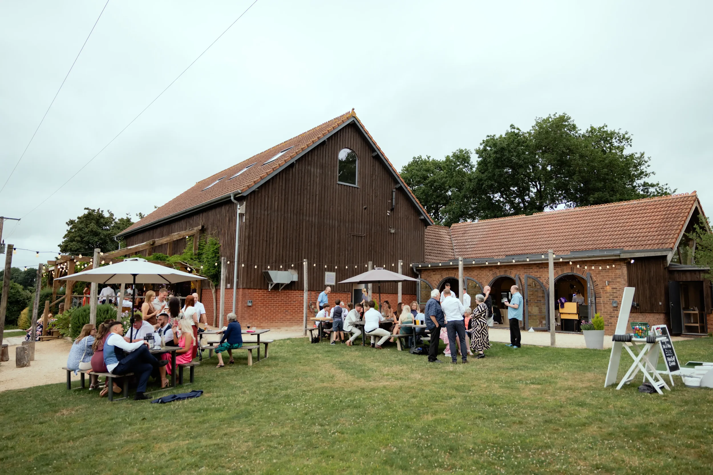 Outdoor gathering at a rustic barn with people sitting at picnic tables and standing, some with drinks, with string lights, umbrellas, and a grassy area