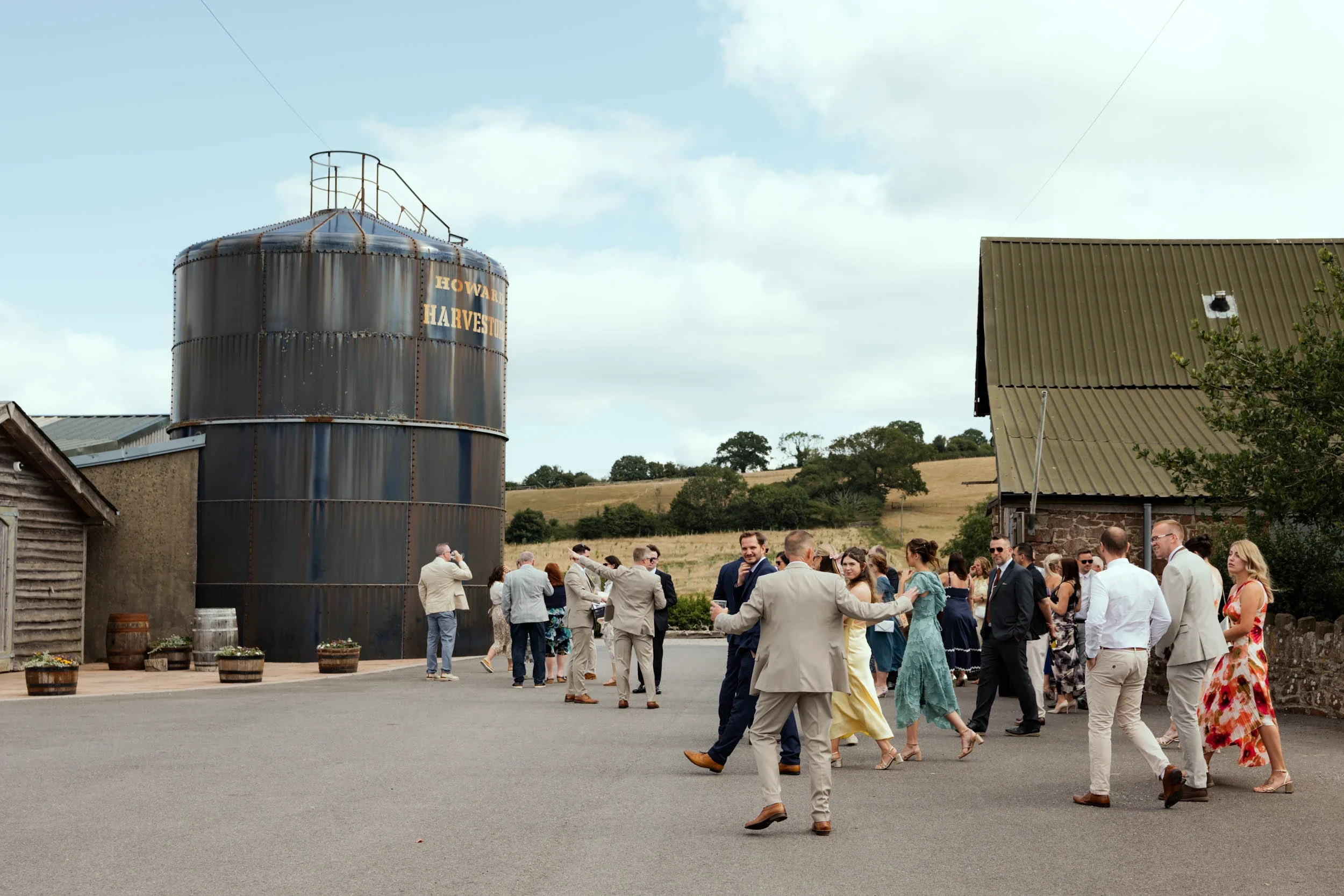 People dressed in formal attire dancing and socializing outdoors near large rusted silo labeled "Howard Harvest" on a farm with rolling hills in the background.