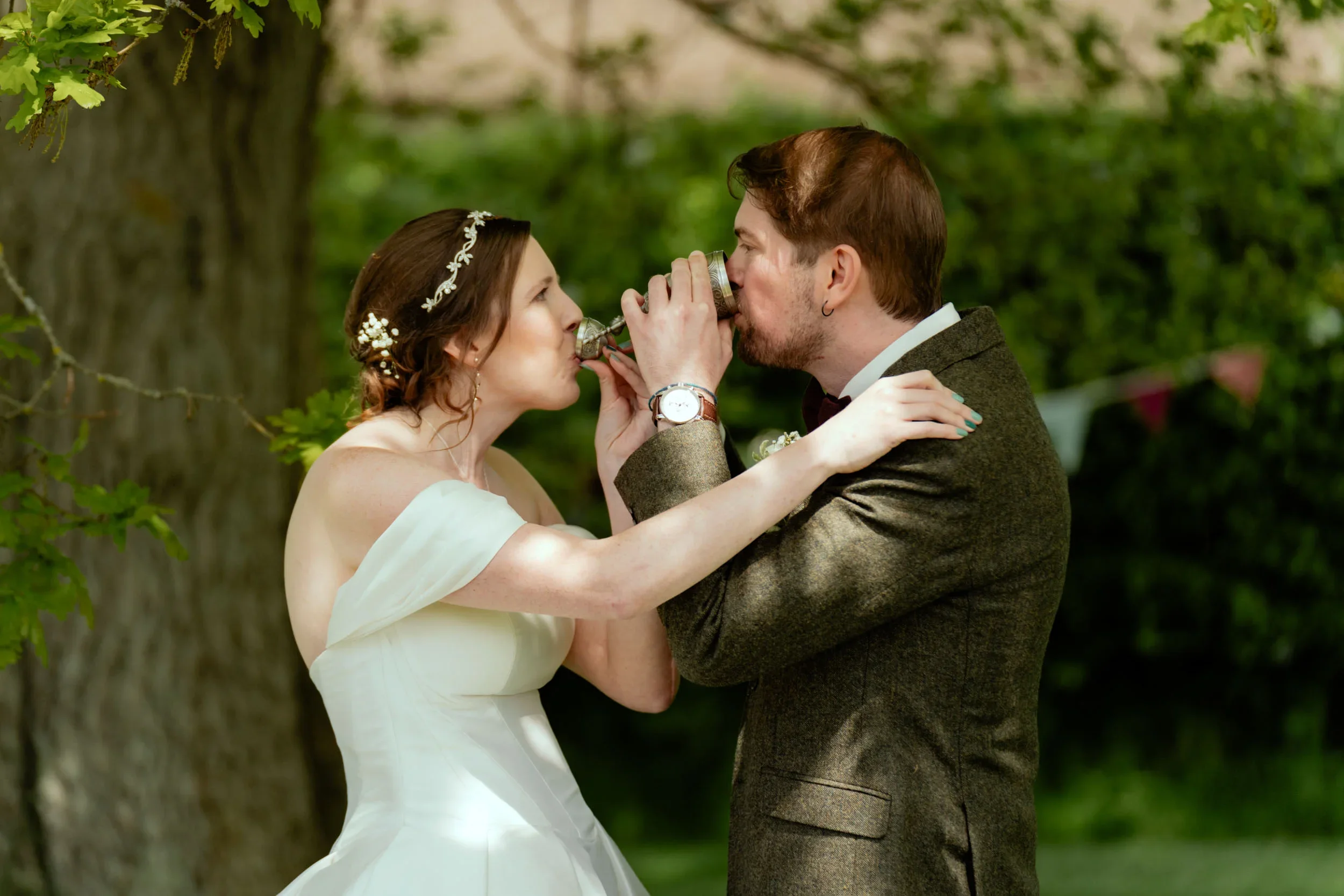 Wedding couple drinking from beer steins outdoors with green trees in background.