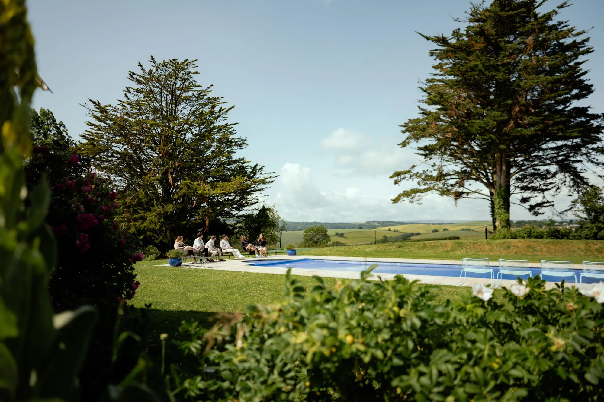 A group of people sitting outdoors near a swimming pool, surrounded by trees and greenery, with a scenic countryside view in the background.