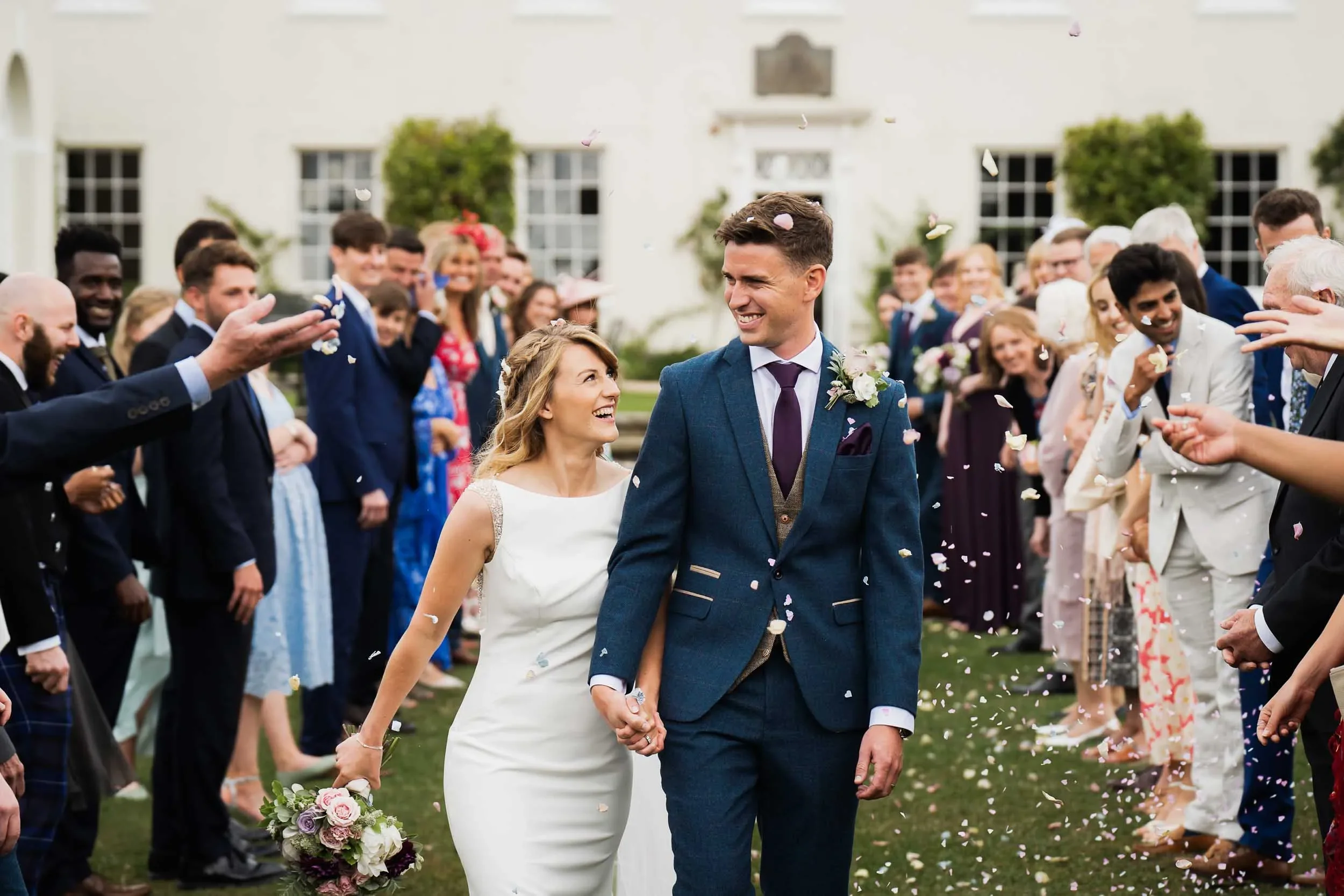 A bride and groom walking hand in hand through a crowd of celebrating wedding guests outside a large white building, surrounded by falling confetti.