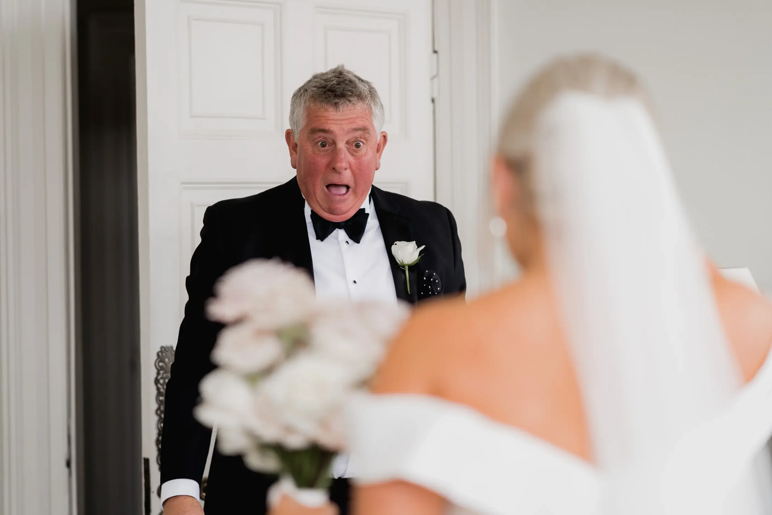 An older man in a tuxedo with a surprised or shocked expression looking at a woman in a wedding dress holding a bouquet of flowers.