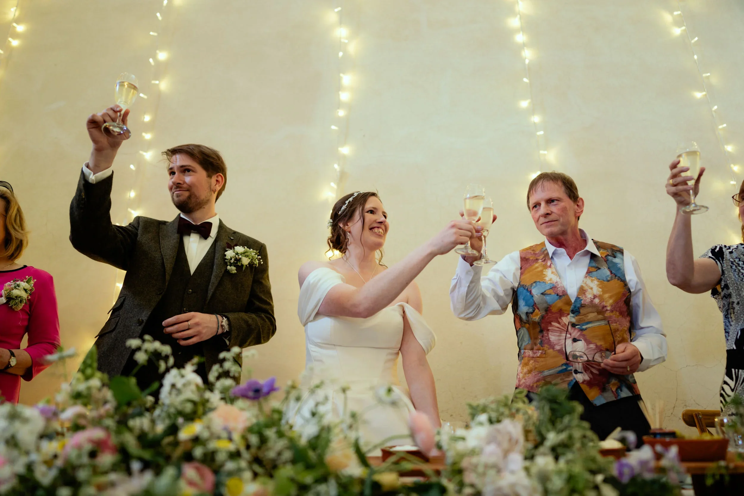 People at a wedding celebration raising glasses of champagne in a toast, smiling and dressed in formal attire, with fairy lights and floral decorations in the background.