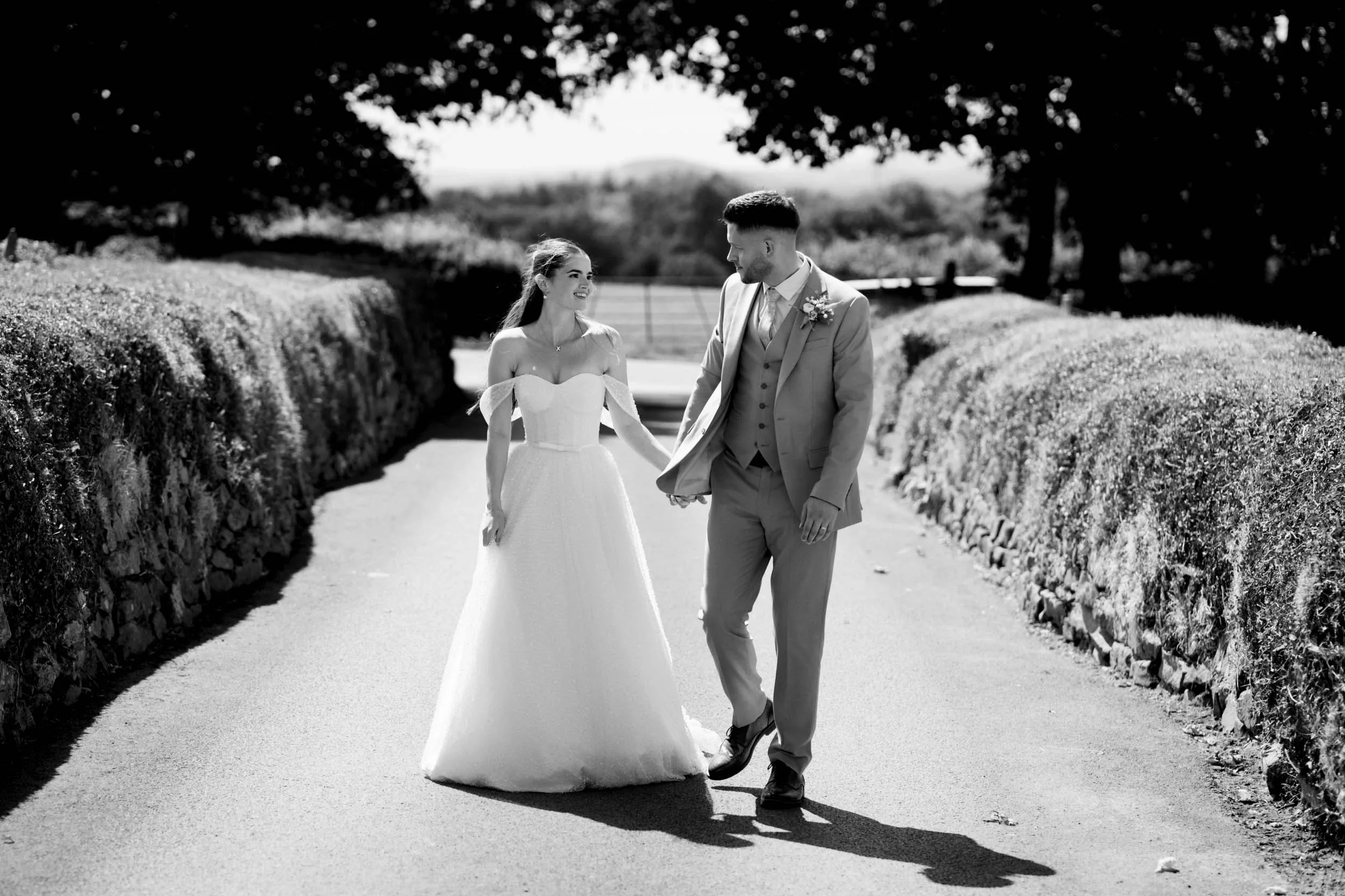 Black and white photo of a bride and groom walking hand in hand outdoors, surrounded by bushes and trees, on a sunny day.