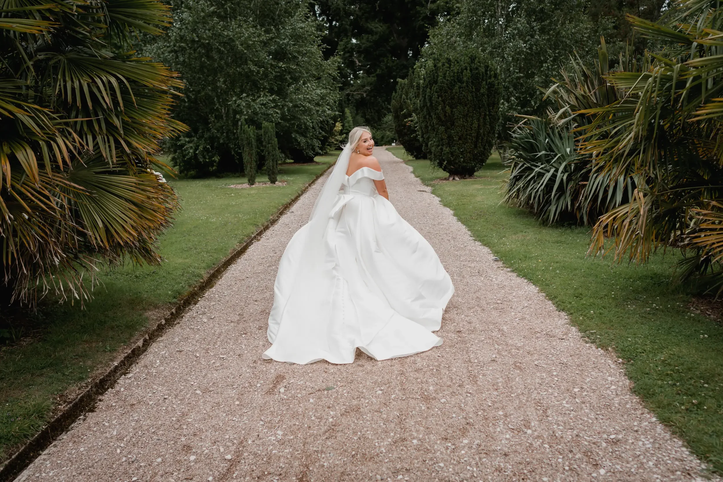 A bride in a white wedding dress and veil smiling and turning on a gravel path surrounded by green trees and bushes.