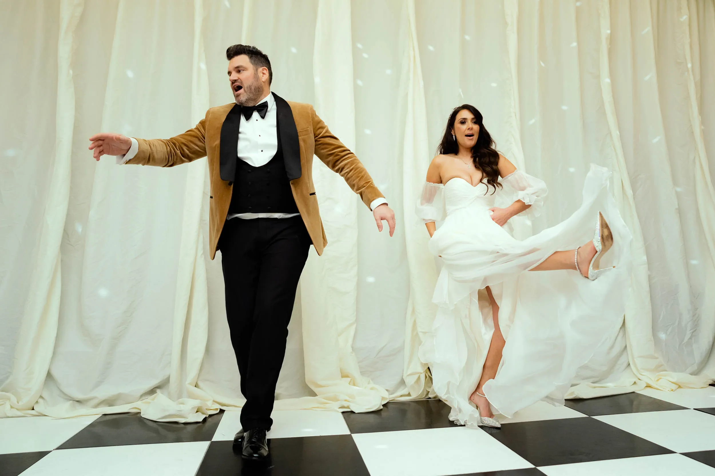 A man and woman dancing at a wedding reception, with the man wearing a brown tuxedo jacket, and the woman in a white wedding dress lifting her leg with silver heels, on a black and white checkered floor with cream curtains in the background.