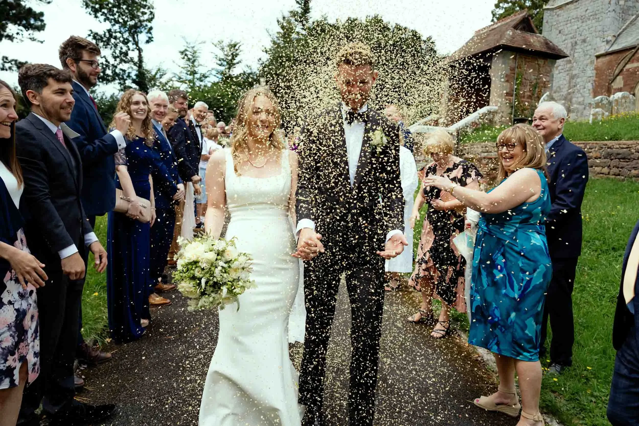 A newlywed couple walking hand in hand on their wedding day, surrounded by friends and family in an outdoor setting, with confetti being thrown in celebration.