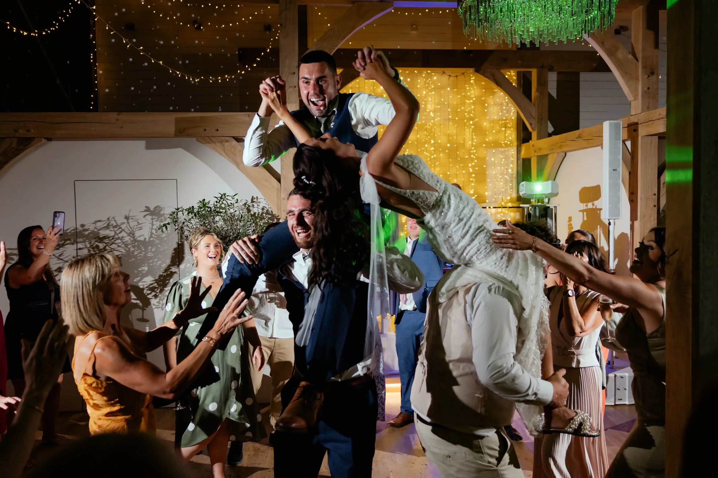 A lively celebration at a wedding reception with people dancing and cheering. Two men are carrying a woman dressed in a white wedding gown on their shoulders, while guests around them are enjoying and taking photos. The scene is decorated with string