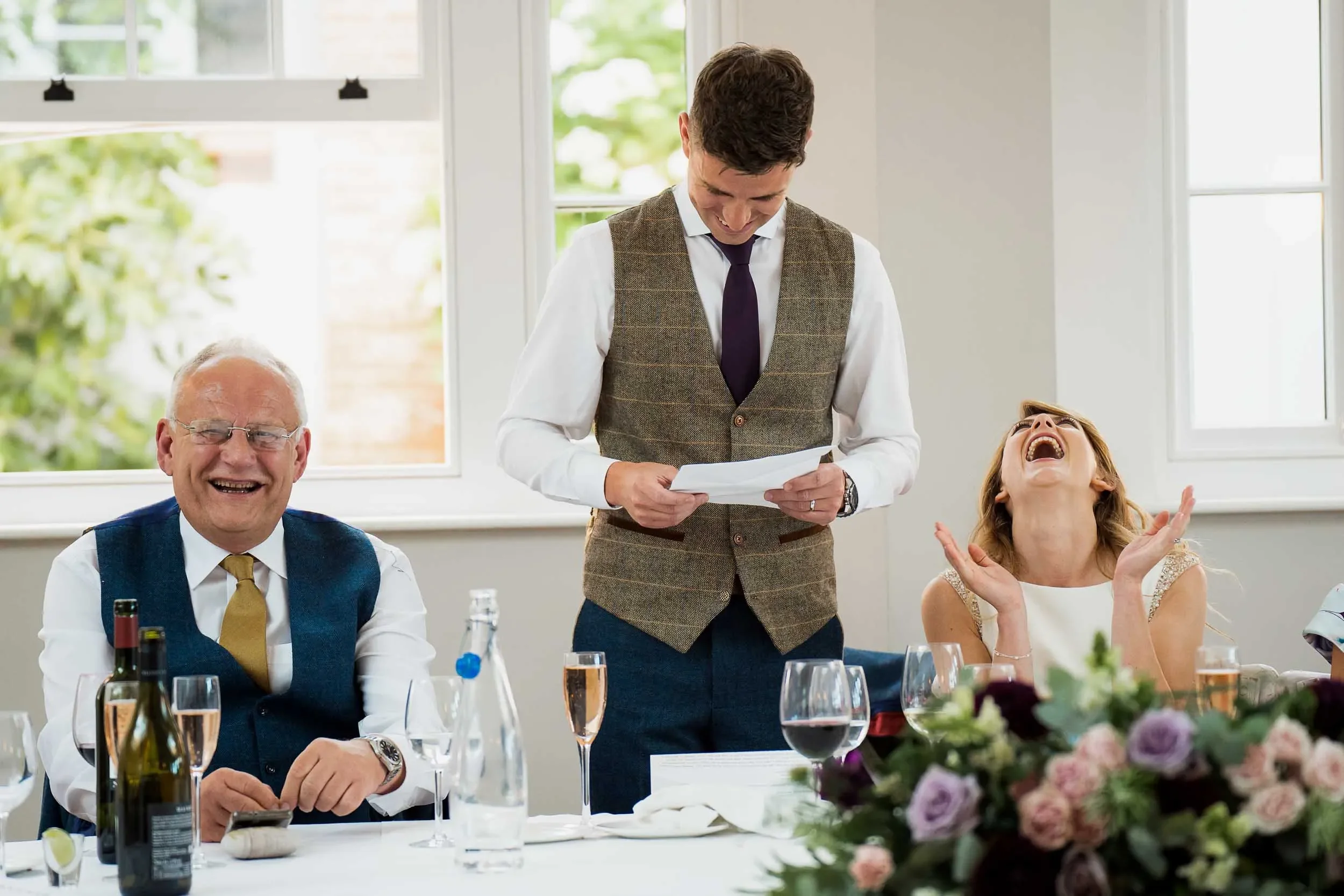 A man standing and smiling while reading a paper during a celebration, with a seated older man and a laughing woman at a table decorated with flowers and drinks.
