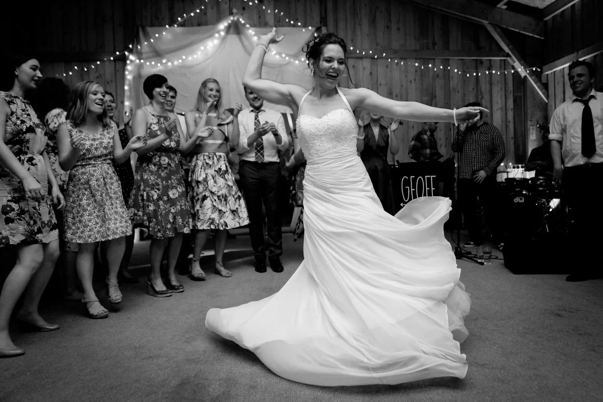 A bride in a wedding dress dancing at her wedding reception, surrounded by guests in a rustic barn setting with string lights.