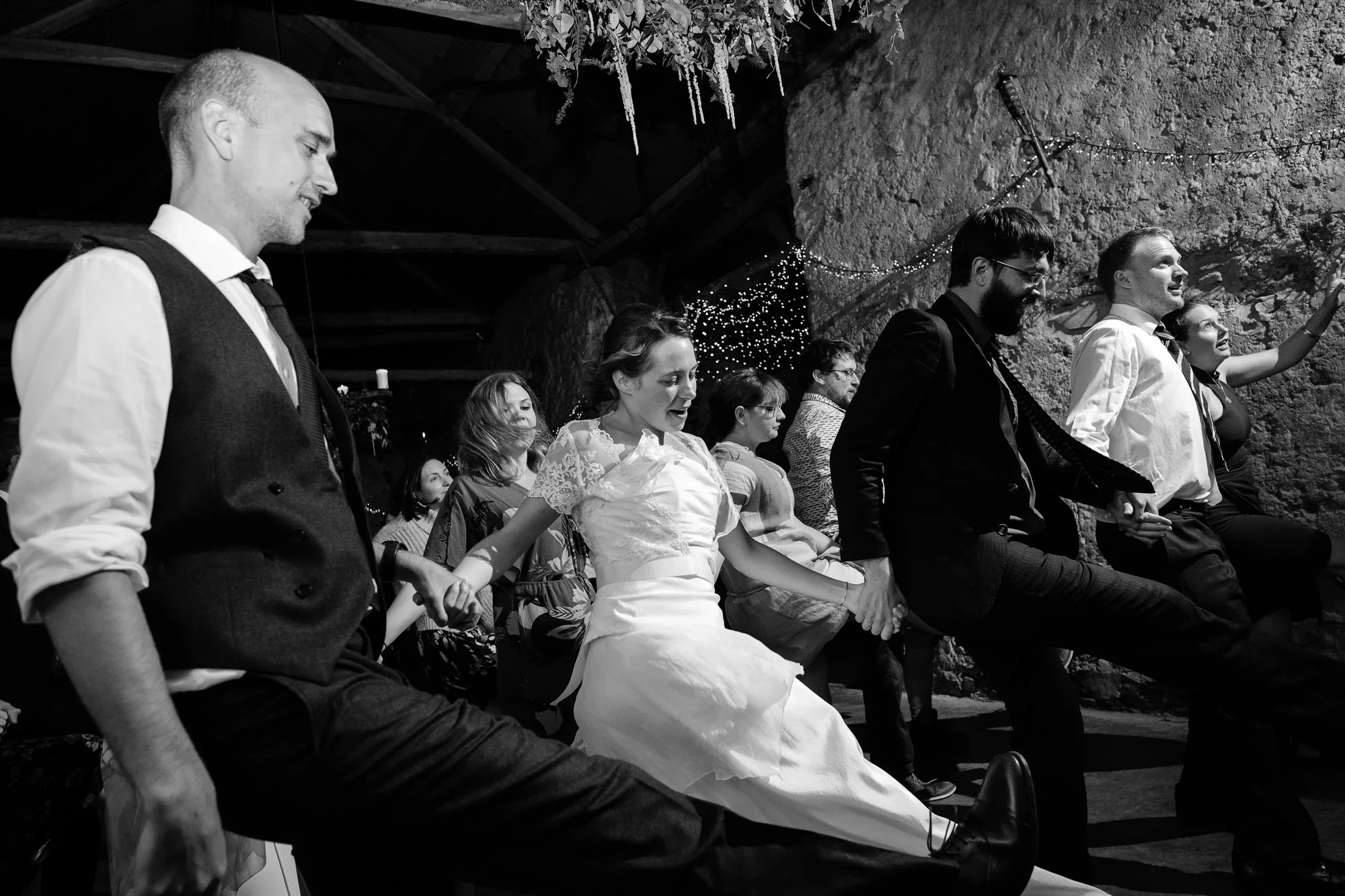 Group of people holding hands and sitting on chairs, participating in a line dance at a celebration or wedding in a rustic venue with string lights and decorations.