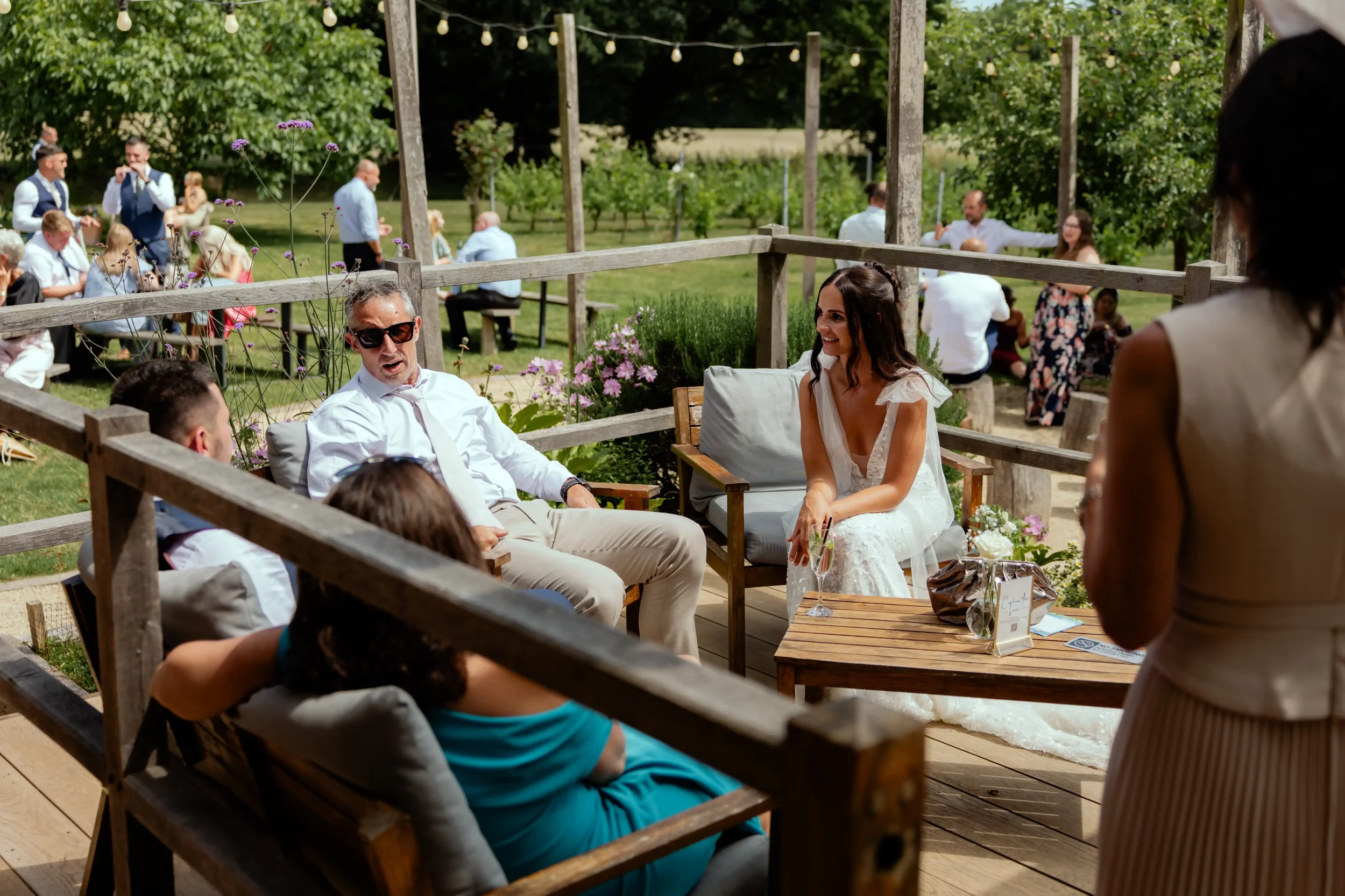 People seated outdoors at a social event, with some in wedding attire, on a wooden deck decorated with flowers and string lights, with a background of green trees and more guests in a park-like setting.