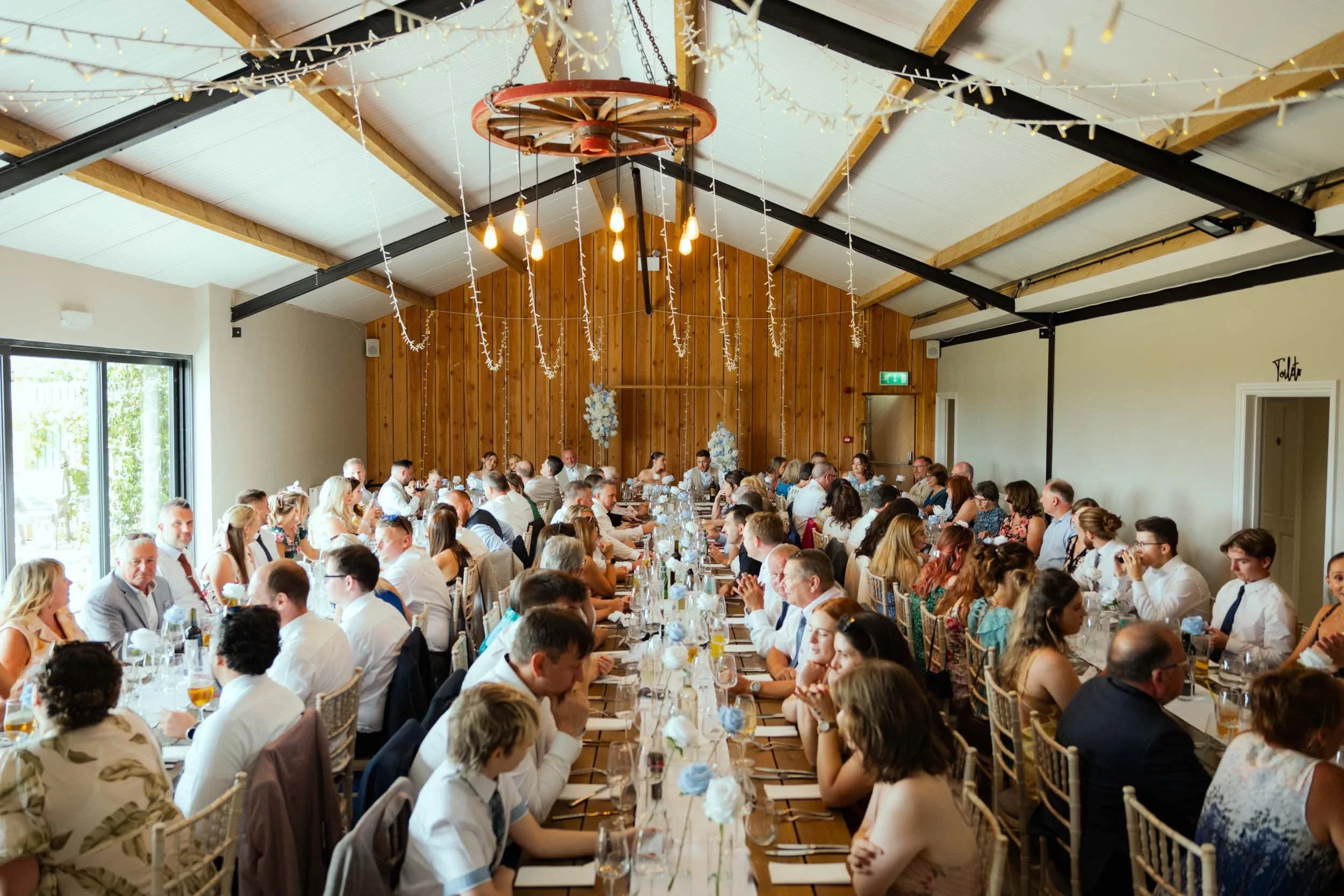 A large gathering of people seated at long banquet tables in a decorated event hall, likely for a wedding or celebration, with wooden wall accents, string lights, and floral centerpieces.