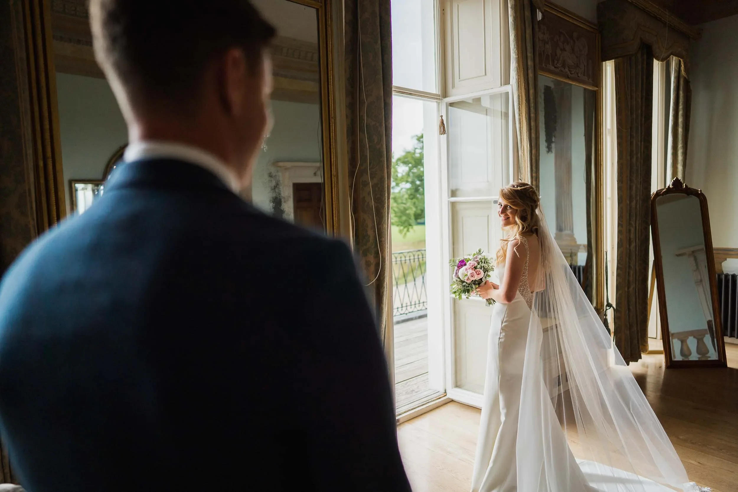 Bride holding a bouquet looking at groom on a wedding day inside a room with large windows and ornate decor.