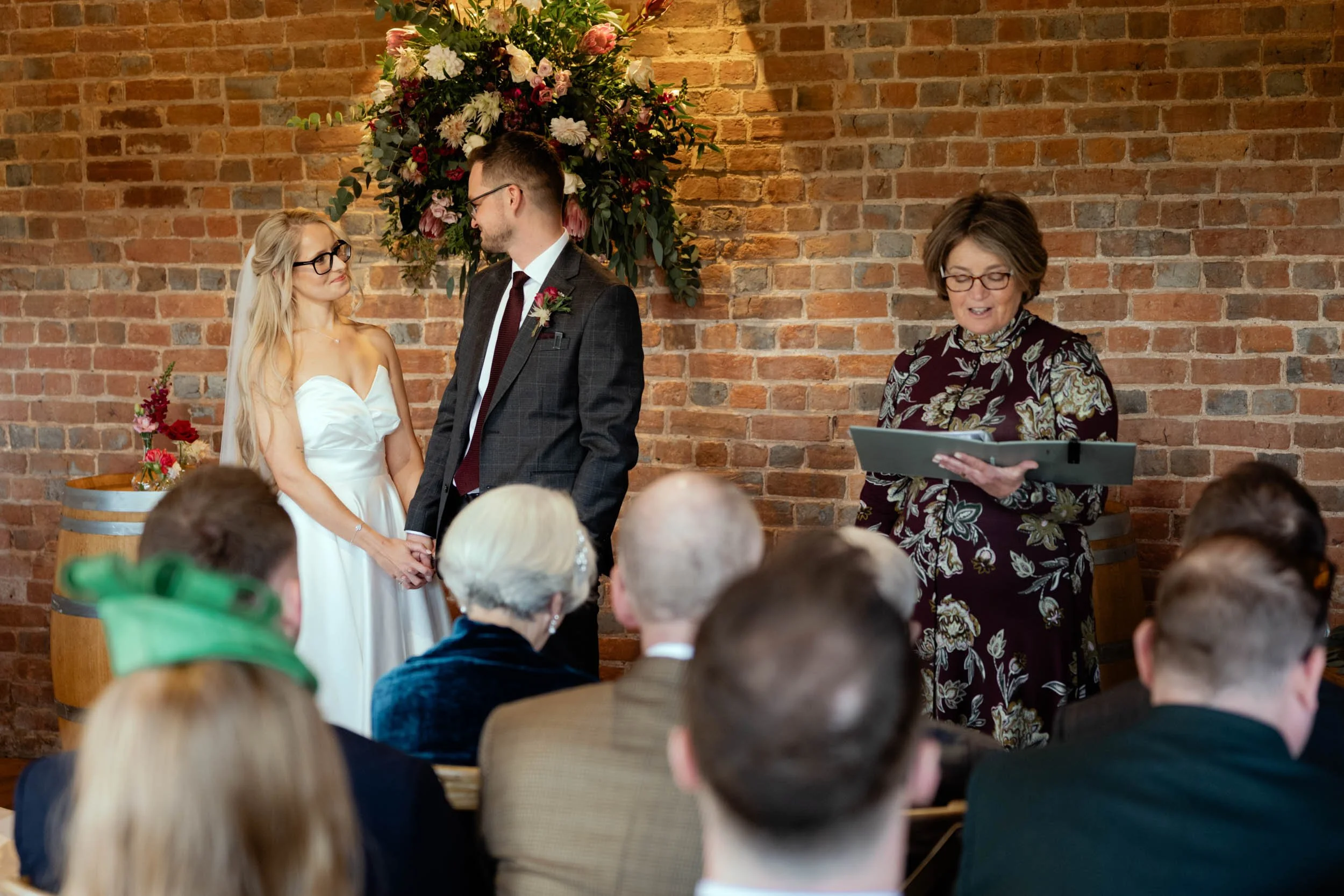 A wedding ceremony with a bride and groom holding hands in front of a brick wall and floral arrangement, officiated by a woman holding a book, with guests seated in front.