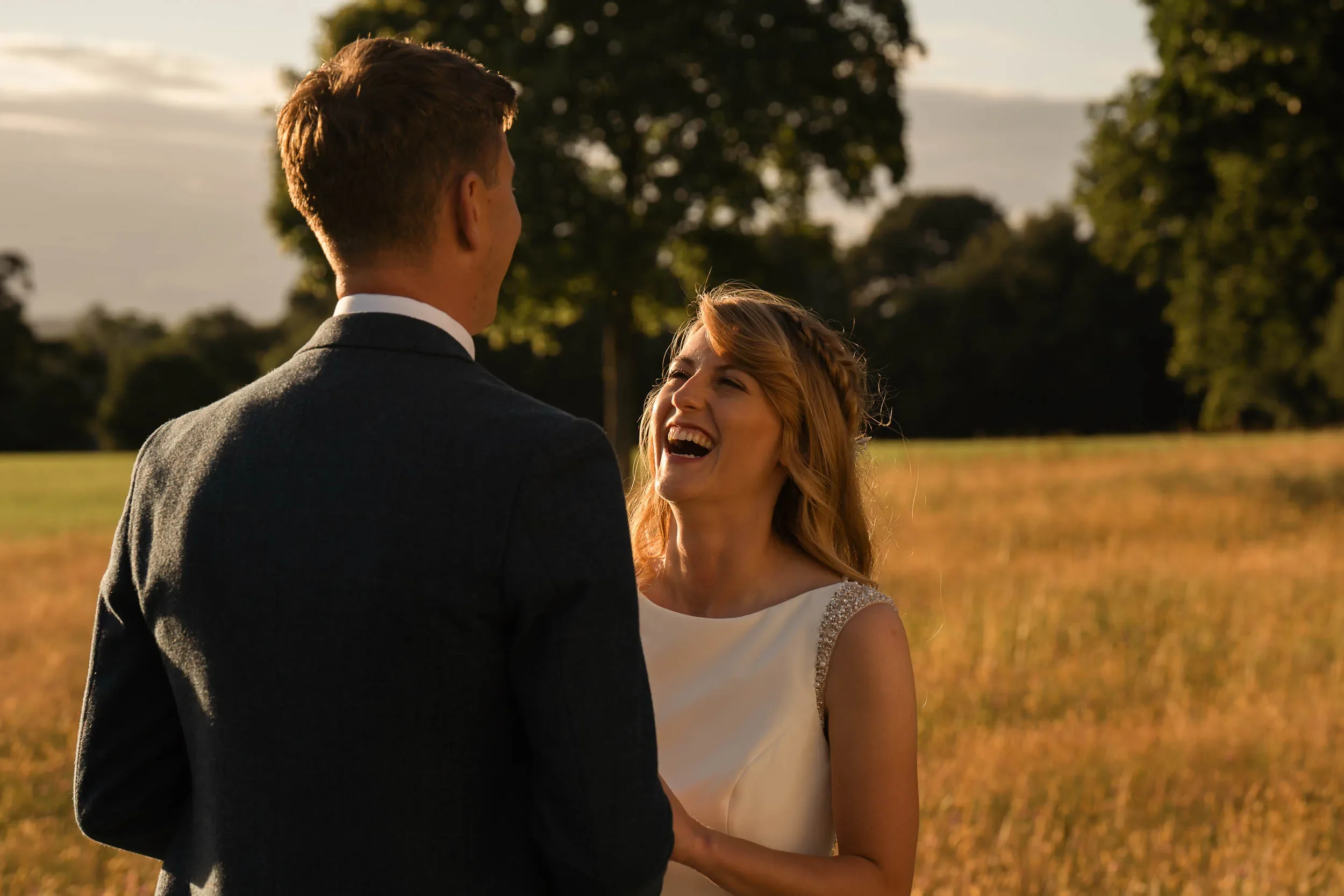 A joyful woman in a white dress with beaded straps laughing and smiling at a man in a suit outdoors during sunset, with a field and trees in the background.