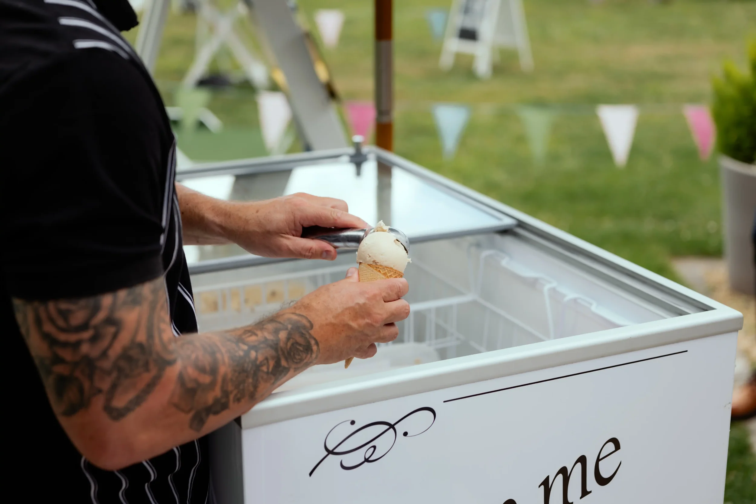 A person with tattooed arms scoops ice cream into a cone from an open freezer at an outdoor event.