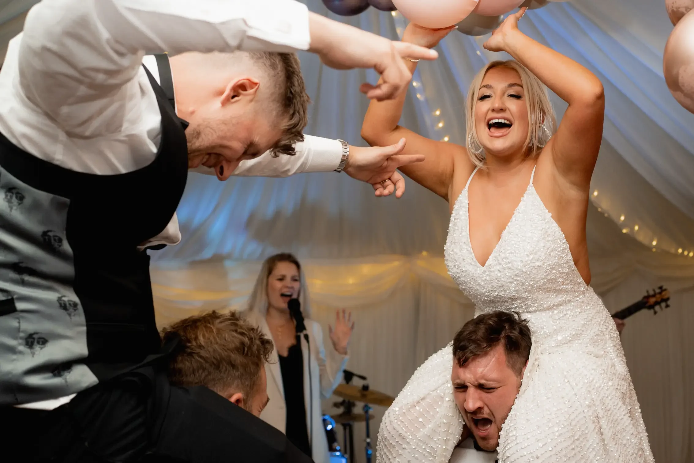 People celebrating at a wedding reception, with a bride in a white dress and a groom, holding a woman on his shoulders, dancing and having fun. A singer is in the background, performing with a microphone.