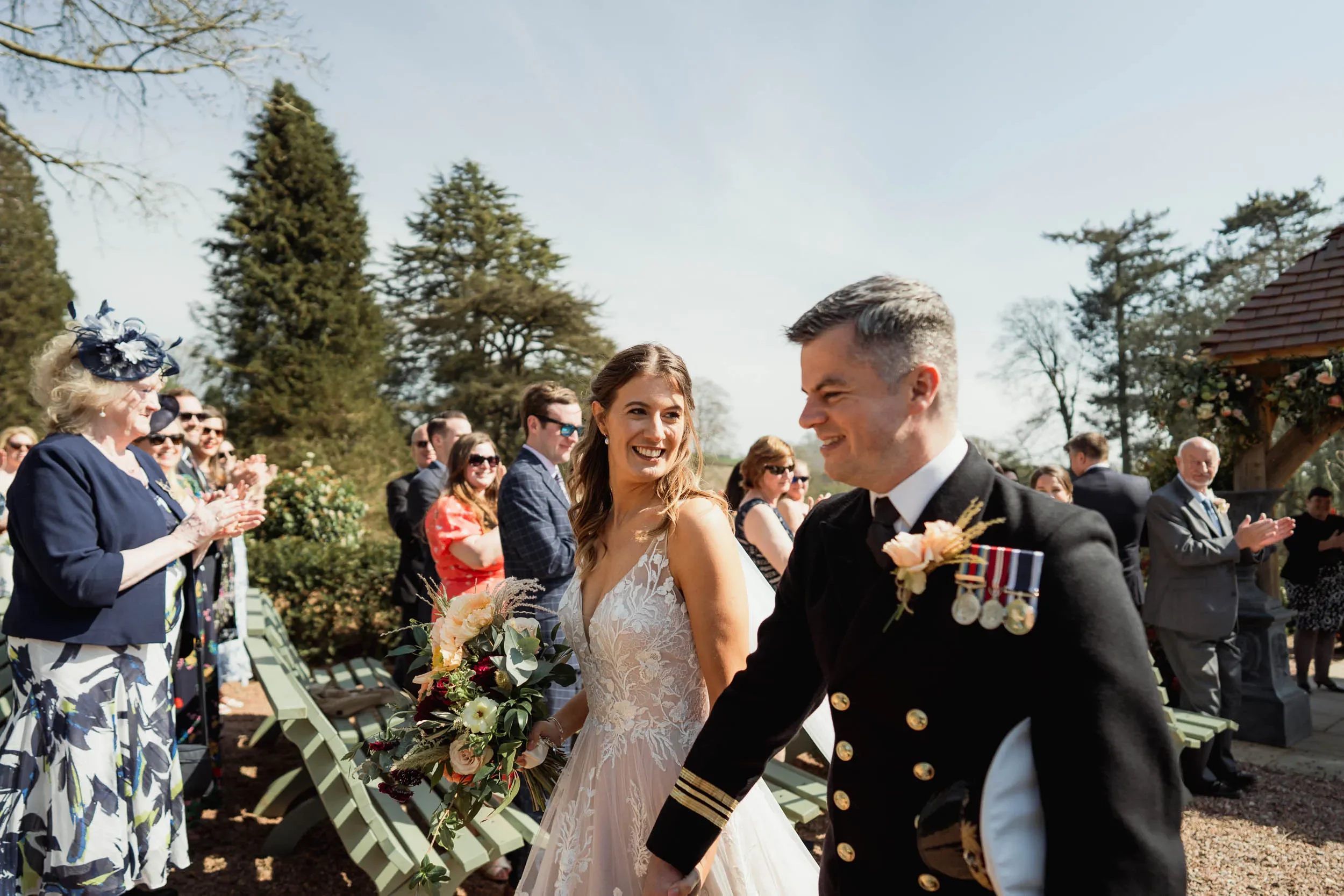 Bride and groom walking hand in hand during a wedding ceremony outdoors, surrounded by guests clapping and smiling.