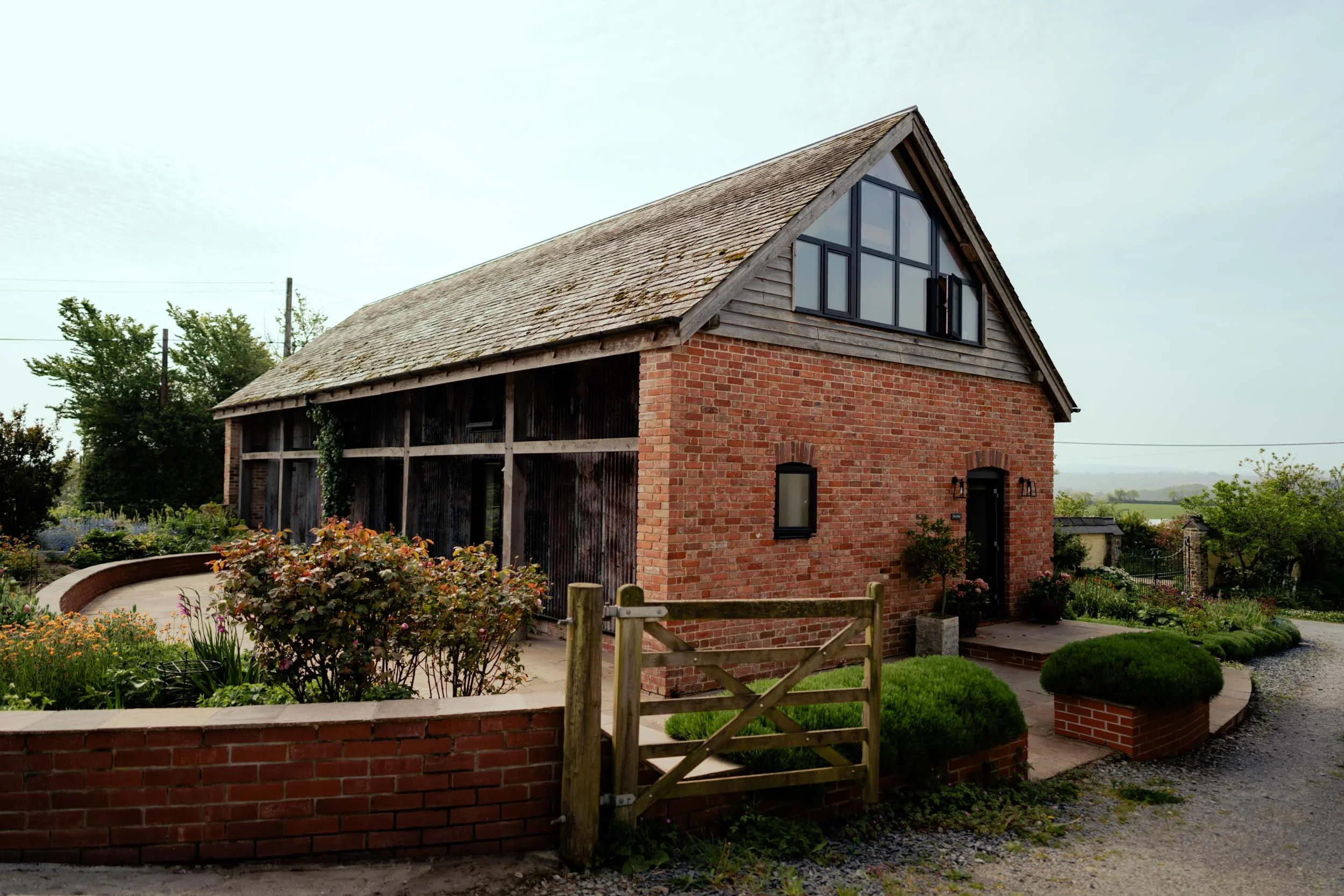 A two-story brick building with a steep roof, large windows on the upper level, and a black front door, surrounded by a landscaped garden with bushes, flowers, and a wooden gate.