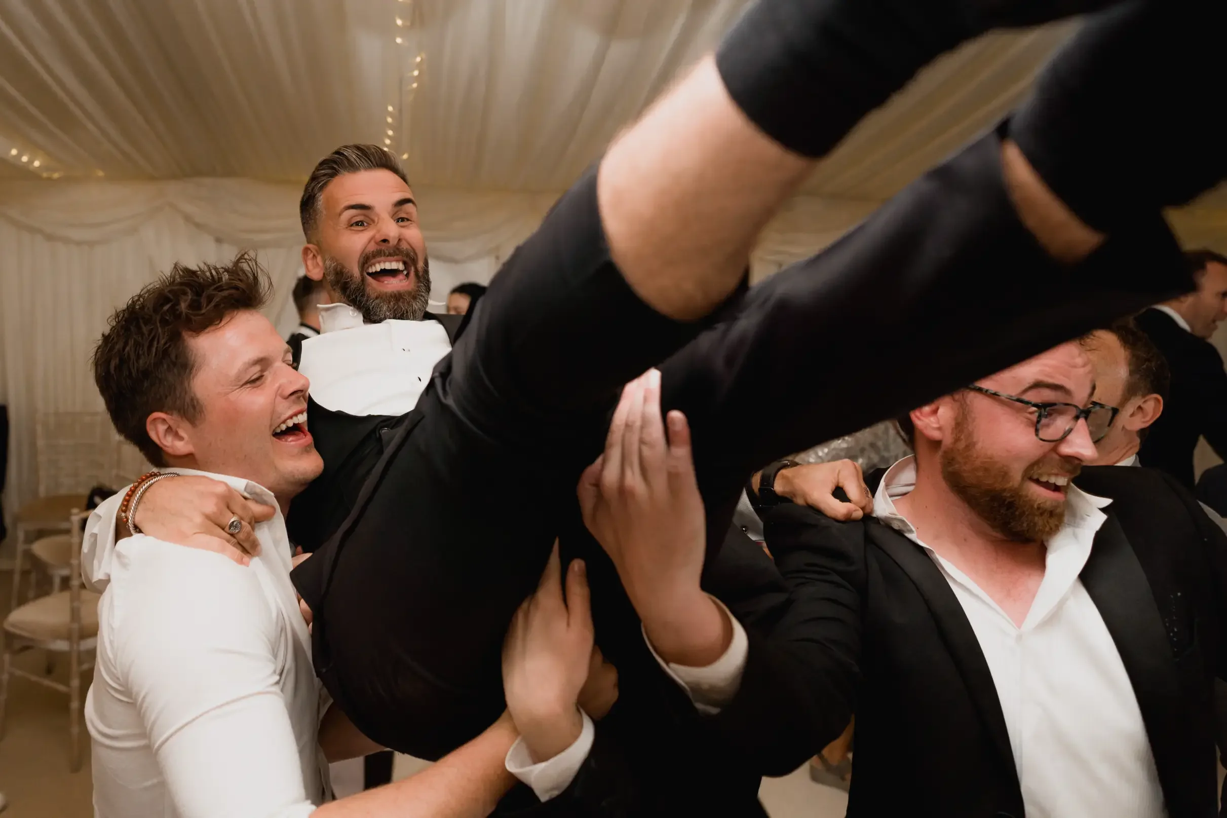 Group of men in formal attire celebrating and lifting a man in a tuxedo at an event under a decorated tent.