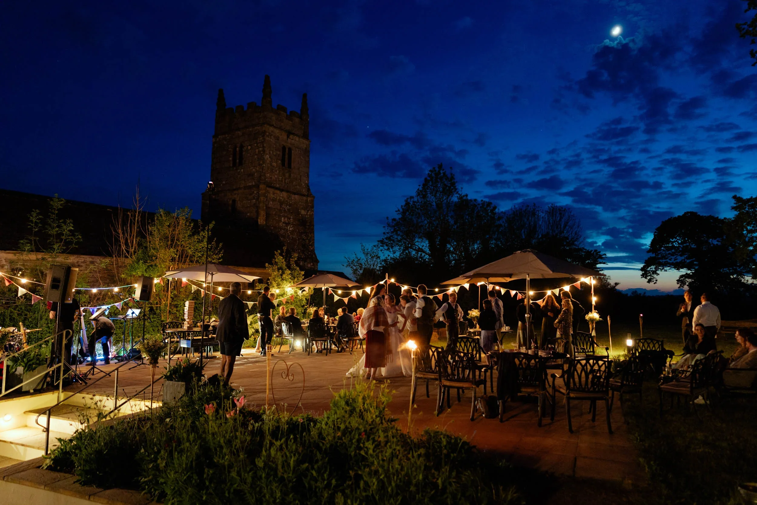 Guests celebrating at an outdoor wedding reception at night, with string lights, a historic stone tower, and a partly cloudy sky with the moon visible.
